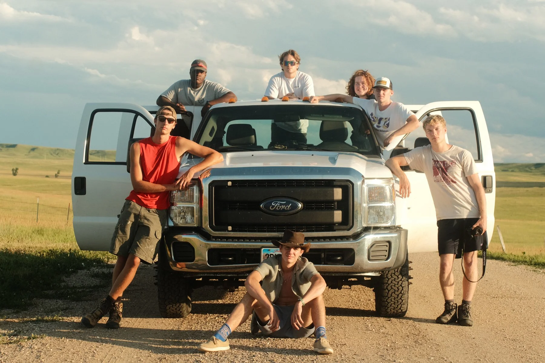 A group of seven young friends with diverse ethnicities, some wearing sunglasses and hats, gather around a white Ford truck in an open countryside landscape with grassy fields and a partly cloudy sky.