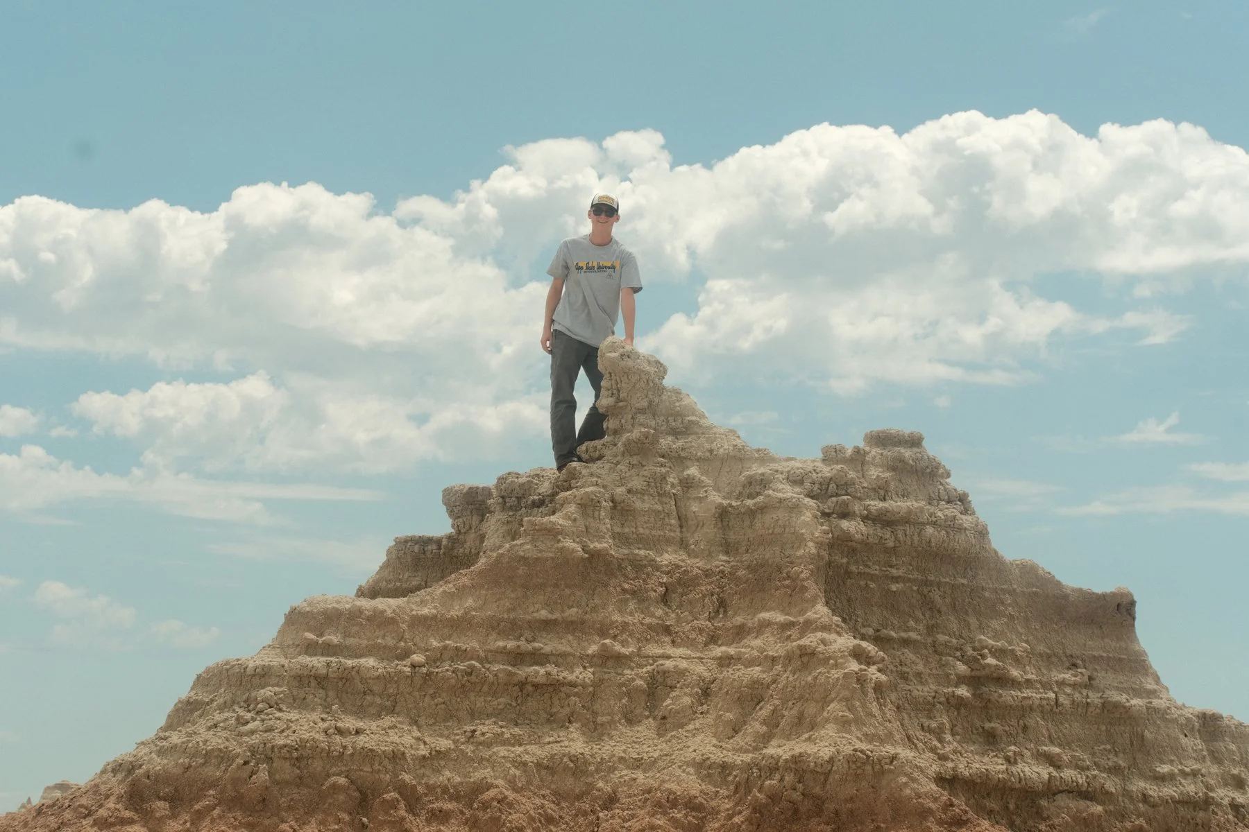 A man standing on top of eroded rock formations in a desert landscape, with a blue sky and scattered clouds in the background.