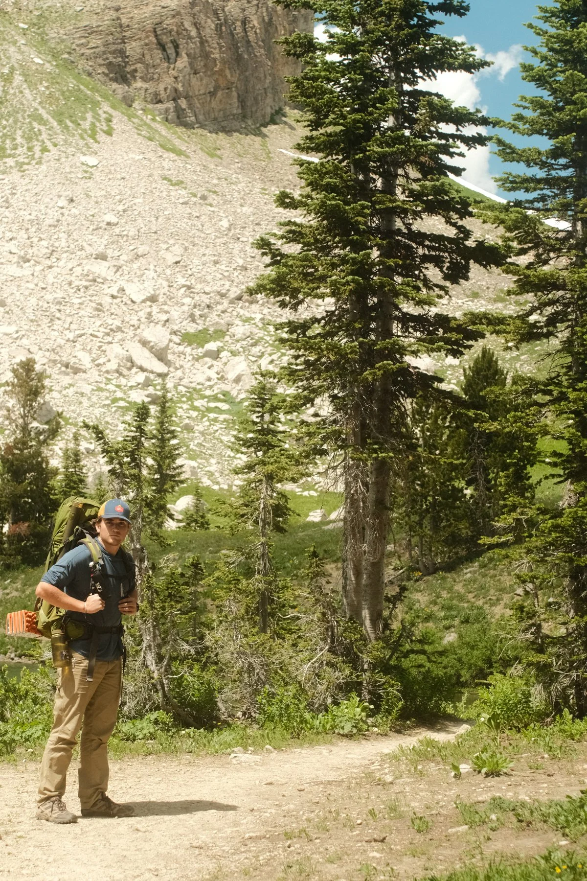 A person hiking in a forested mountain area with tall pine trees, rocky terrain, and a clear blue sky.