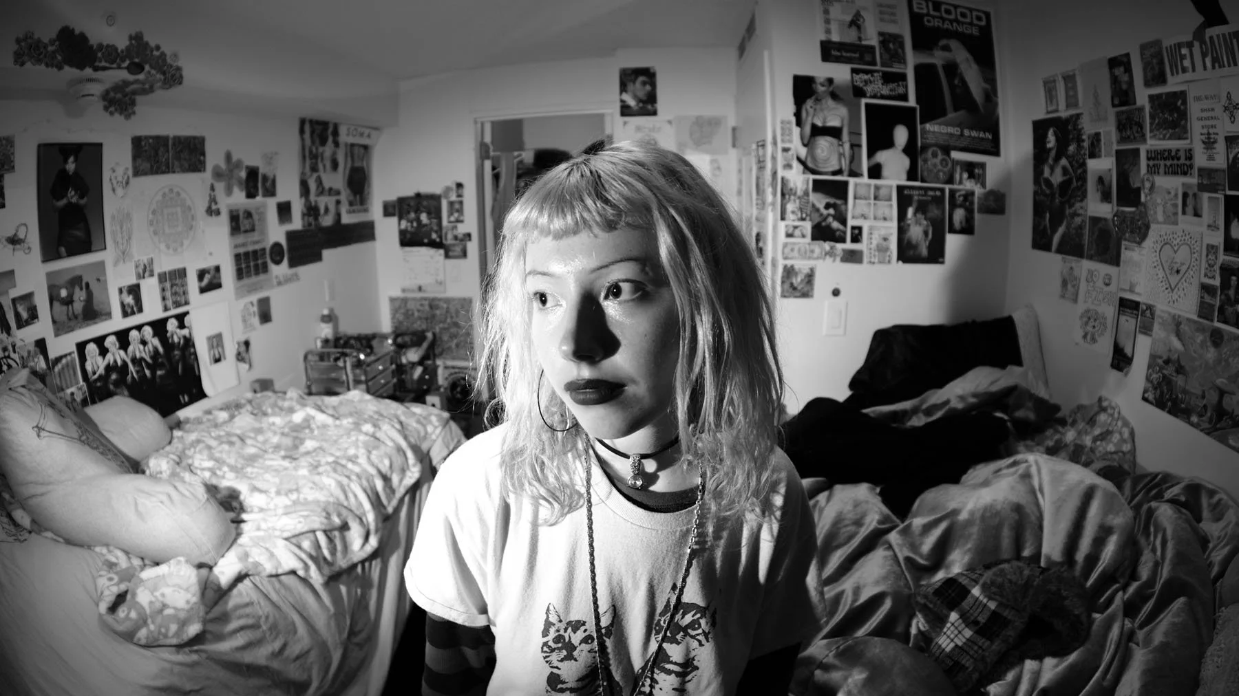 A young woman with light-colored, wavy hair and dark lipstick stands in a cluttered bedroom, surrounded by posters, photos, and artwork on the walls. The bed behind her is unmade with a stuffed animal on it, and the room appears to be decorated with 
