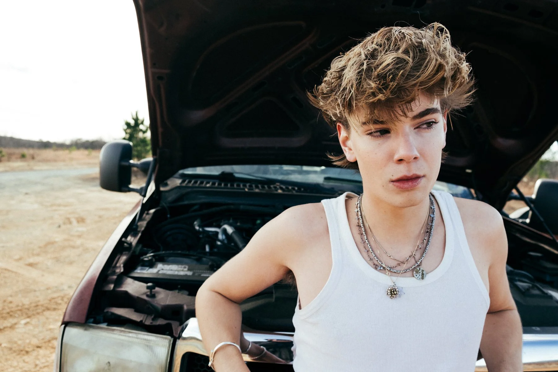 A young person with short, tousled hair and a white tank top standing in front of an open car hood on a dirt road, with a distant landscape and few trees visible in the background.