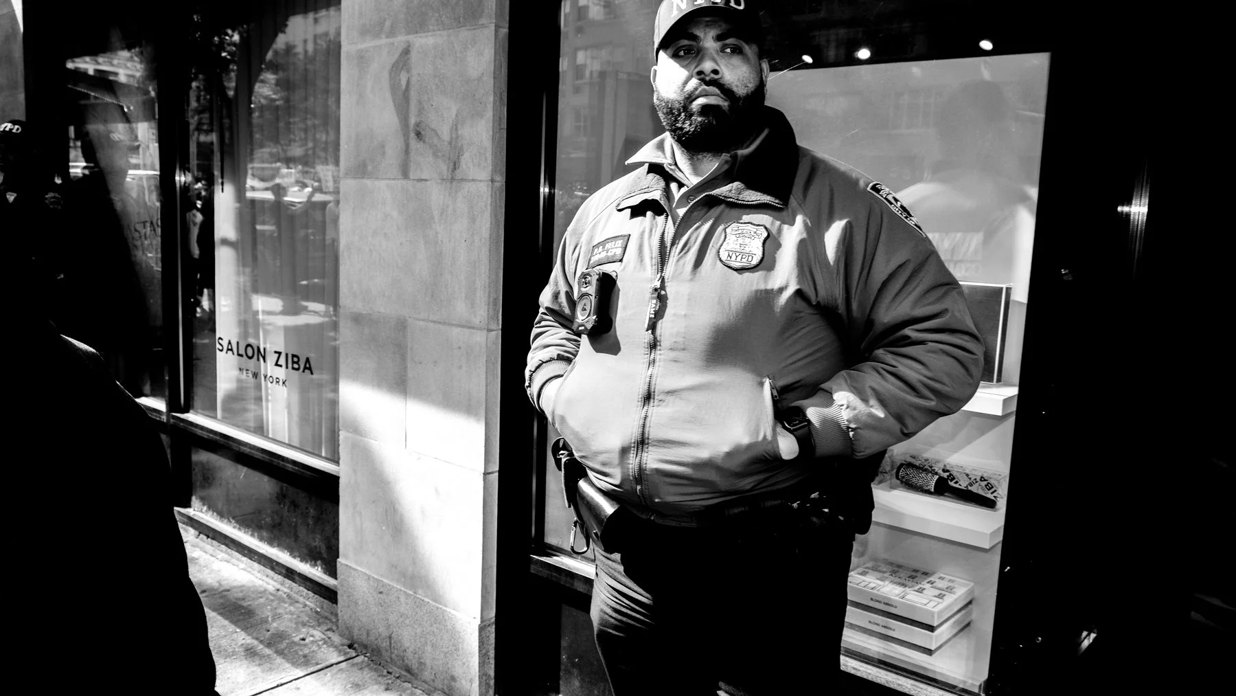 A police officer standing on a city sidewalk in front of a store window, wearing a NYPD jacket and cap, with hands in pockets. Reflection of street scene visible in window, black and white photo.