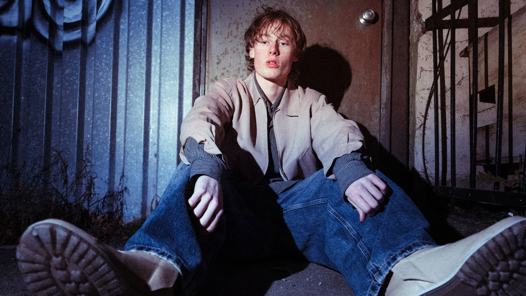 A young man with red hair sitting on the ground in an alleyway, leaning against a wall, with a distressed expression, wearing a beige jacket, blue jeans, and tan boots.