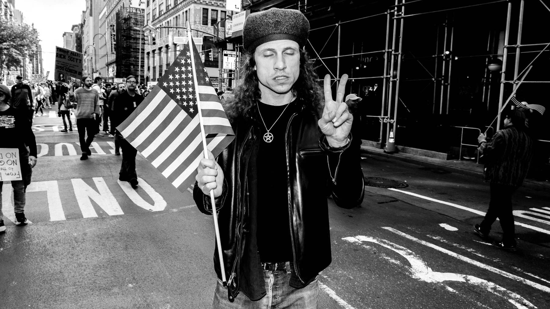A man with long hair wearing a beanie and leather jacket holding an American flag in a protest or rally on a city street, making a peace sign with his other hand. Several people are walking behind him, some holding signs, in an urban setting with tal
