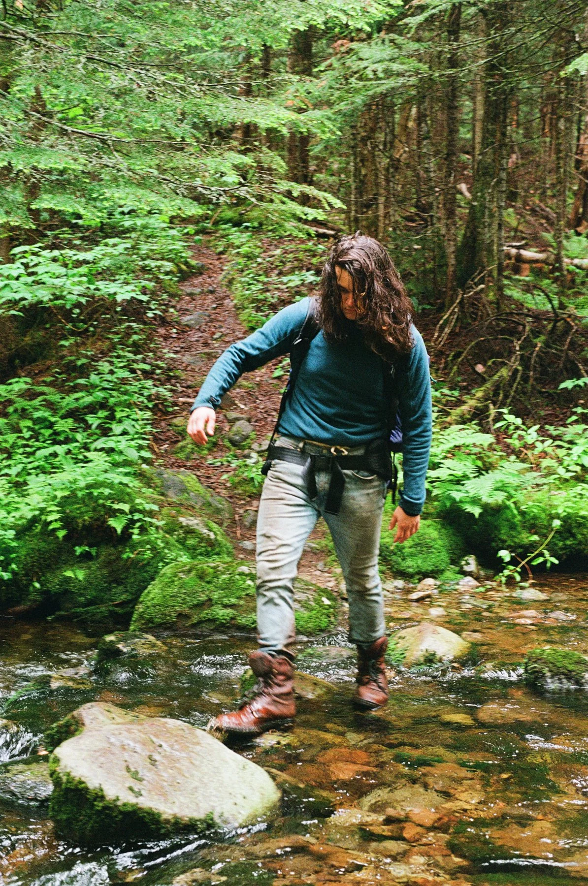 A person with curly hair, wearing a blue jacket, gray pants, and brown boots, crossing a small stream in a lush green forest.