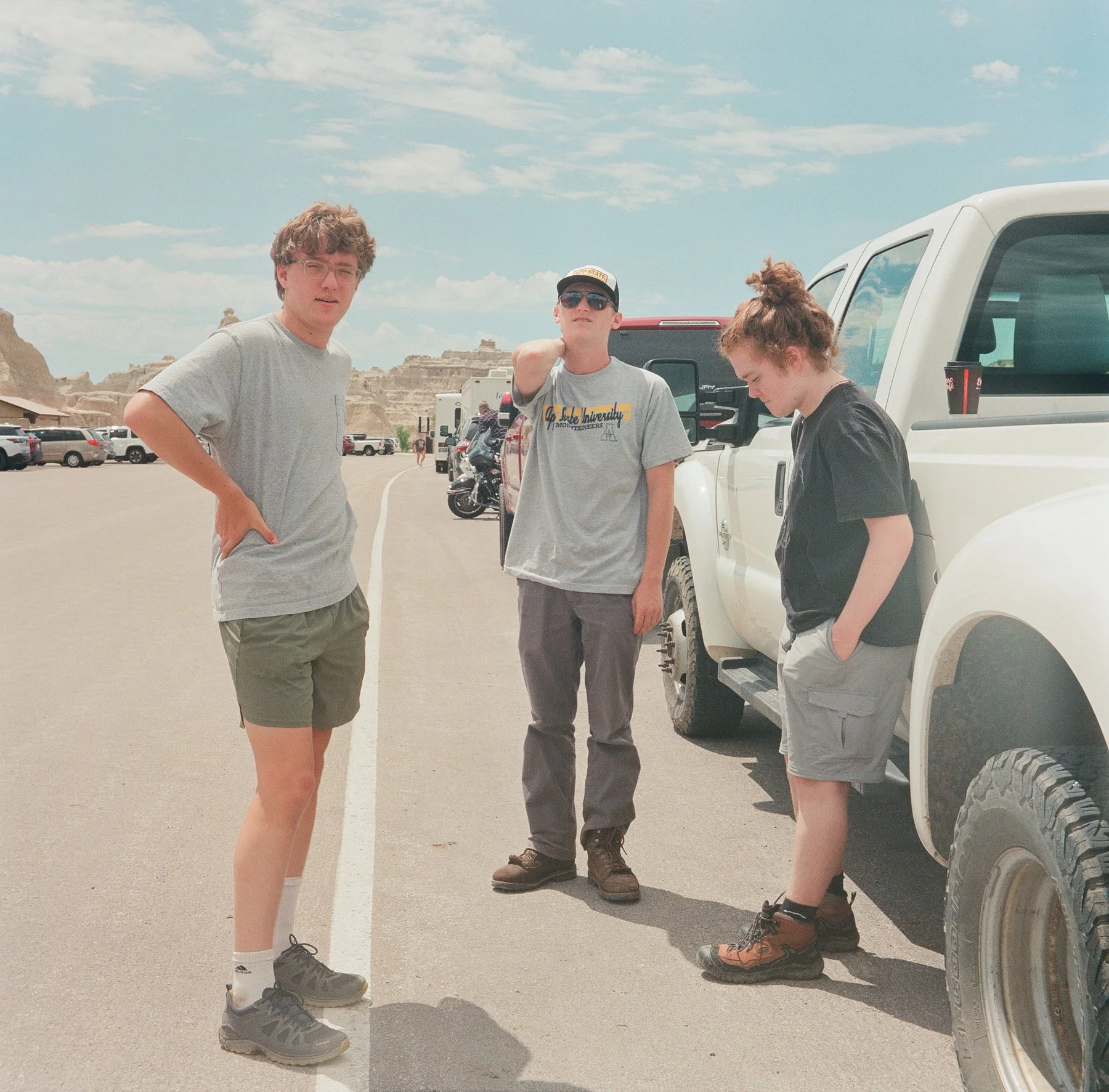 Three young people standing next to a white pickup truck in a desert area with rocky formations in the background under a partly cloudy sky.