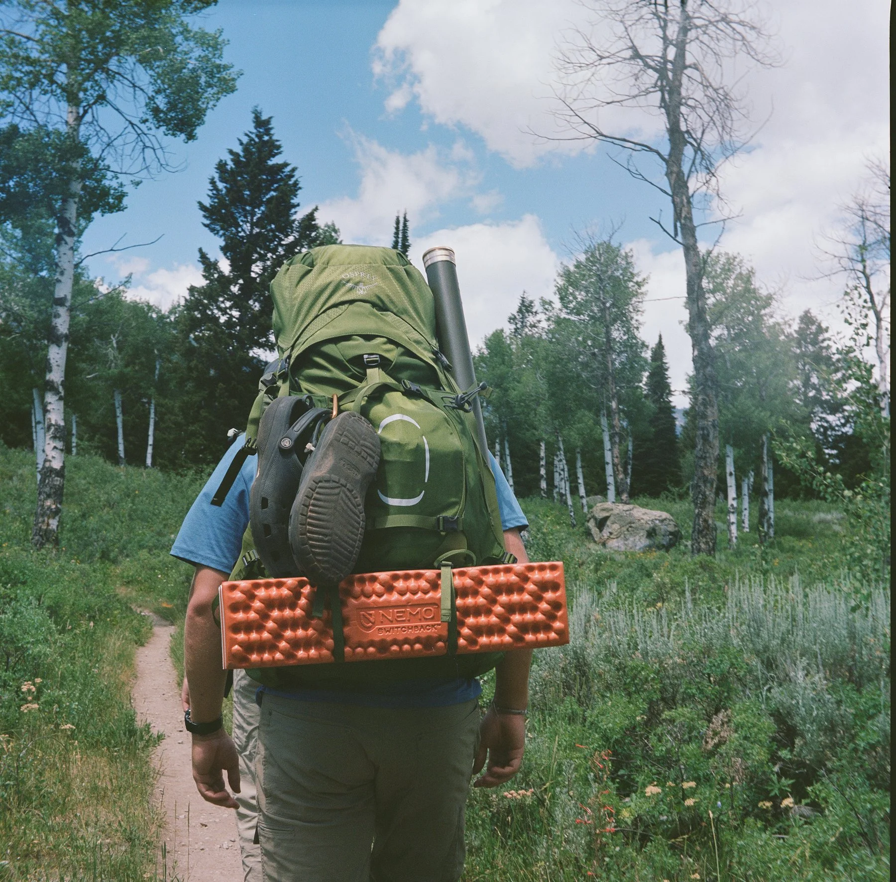 A hiker walking on a forest trail with a large green backpack, a water bottle, and a foam sleeping pad attached. The area is lush with trees and greenery, under a partly cloudy sky.