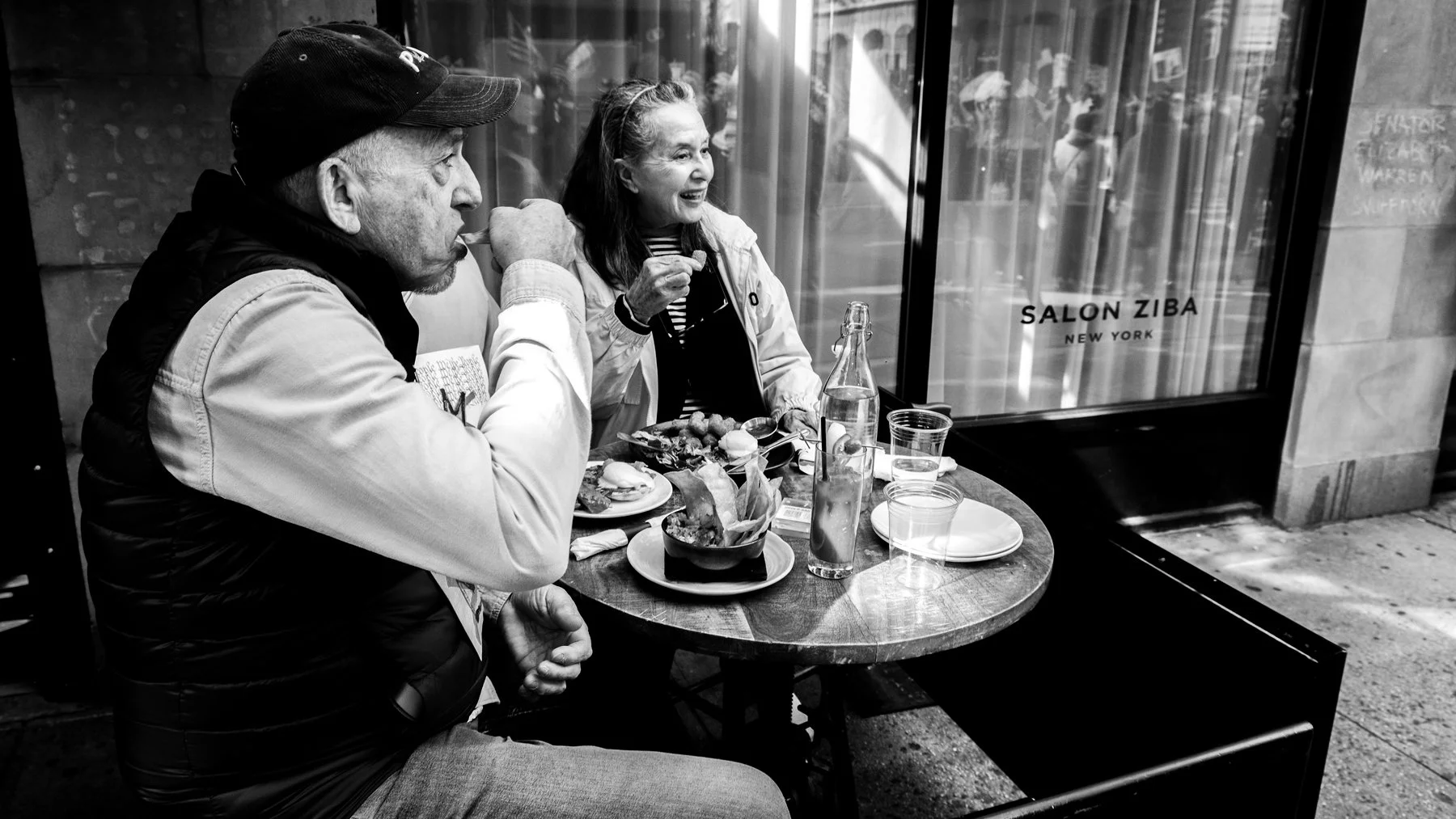 A man and woman sitting at an outdoor café table enjoying a meal in front of a window with the sign 'Salon Ziba New York'.