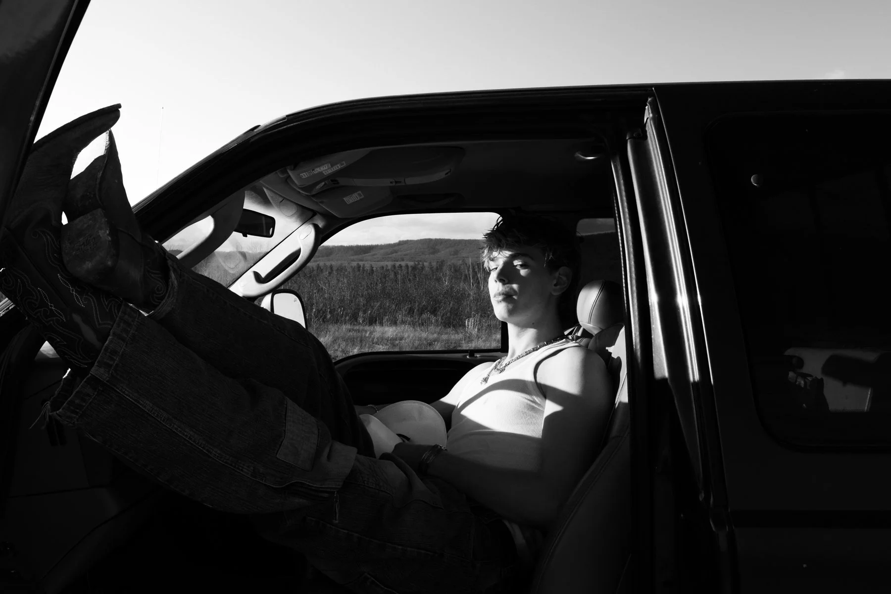 A young person sitting in the driver's seat of a vehicle with the door open, against a landscape background, in black and white.