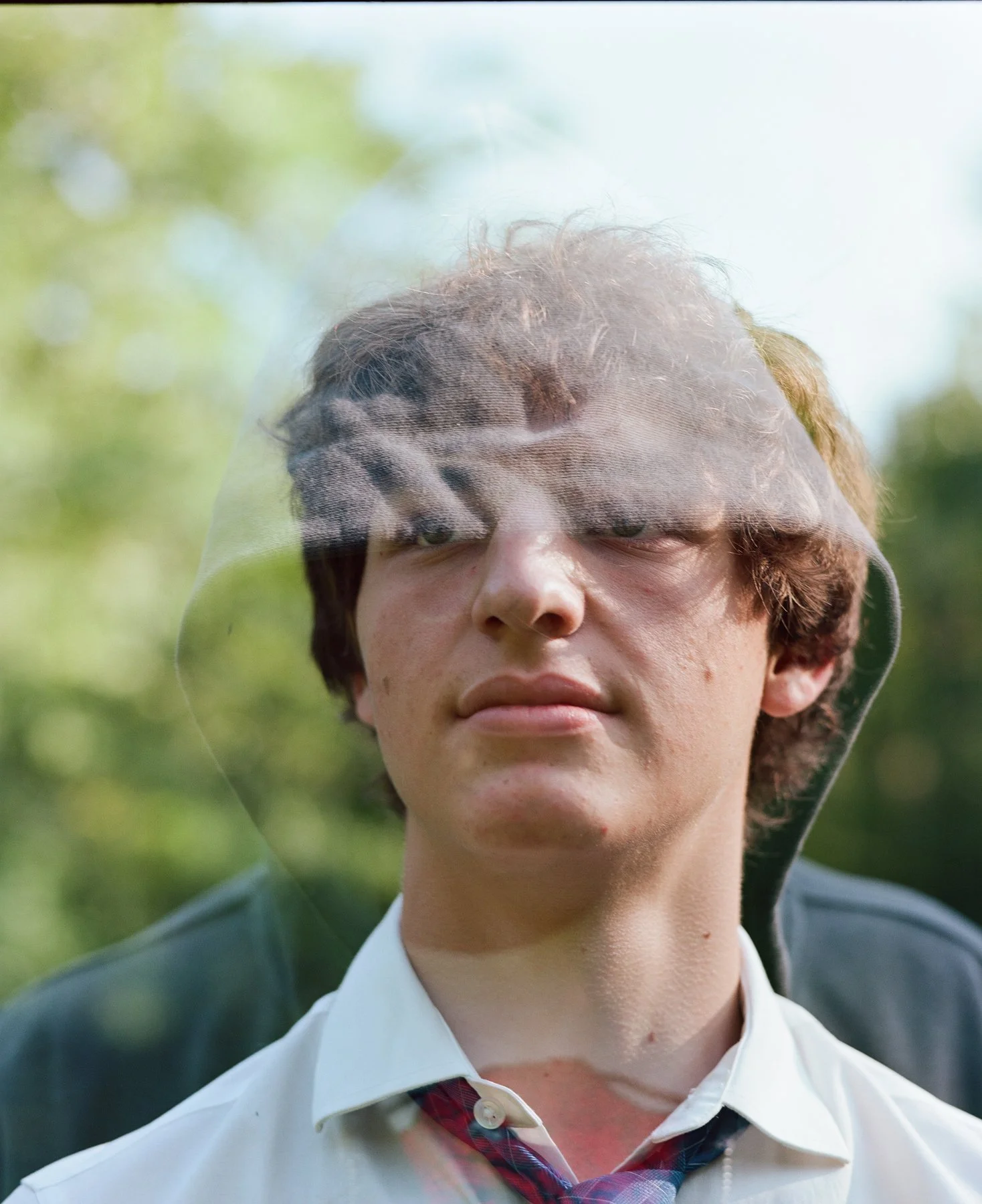 A young man with short brown hair, wearing a white shirt and a loosened plaid tie, is seen through a glass reflection that partially distorts his face. The background shows greenery.