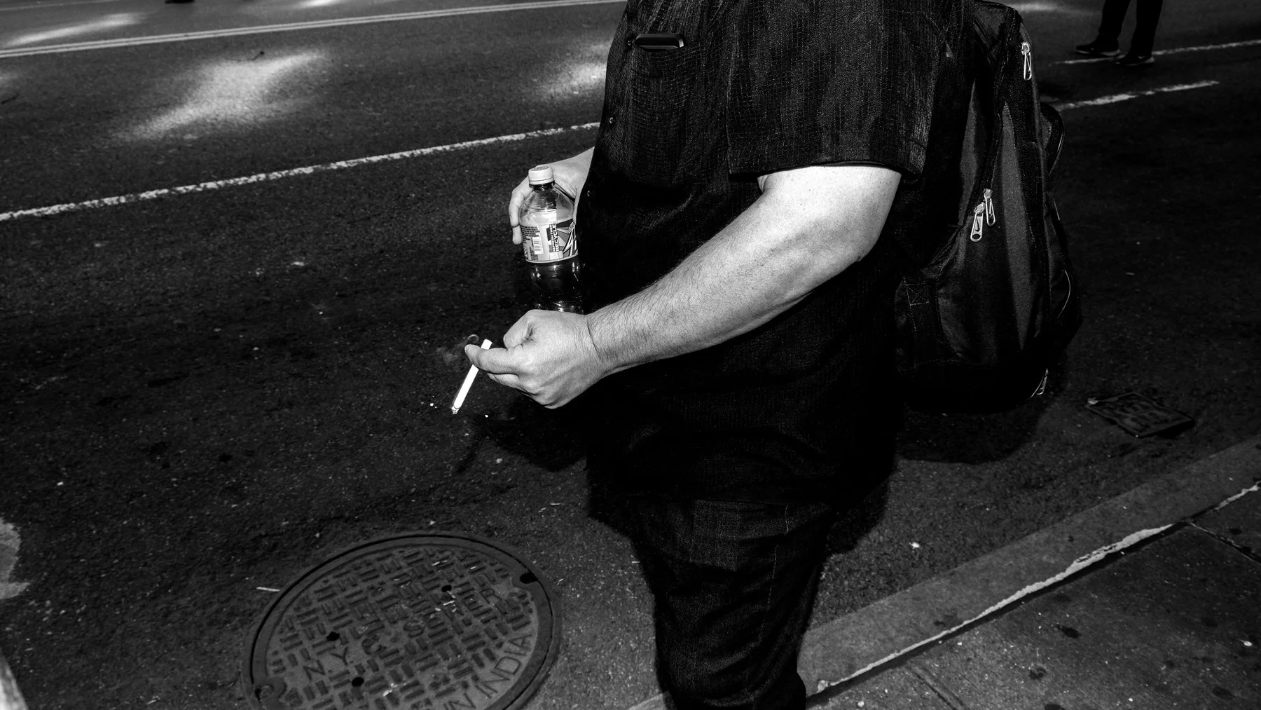 Person holding a cigarette in one hand and a bottle of soda in the other, standing on a city sidewalk at night with a backpack on their back.