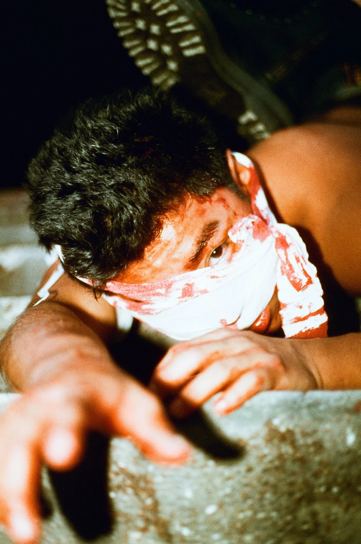 A young man with dark curly hair, wearing a blood-stained bandana over his face and a mask, crawling on the ground, looking tense and focused.