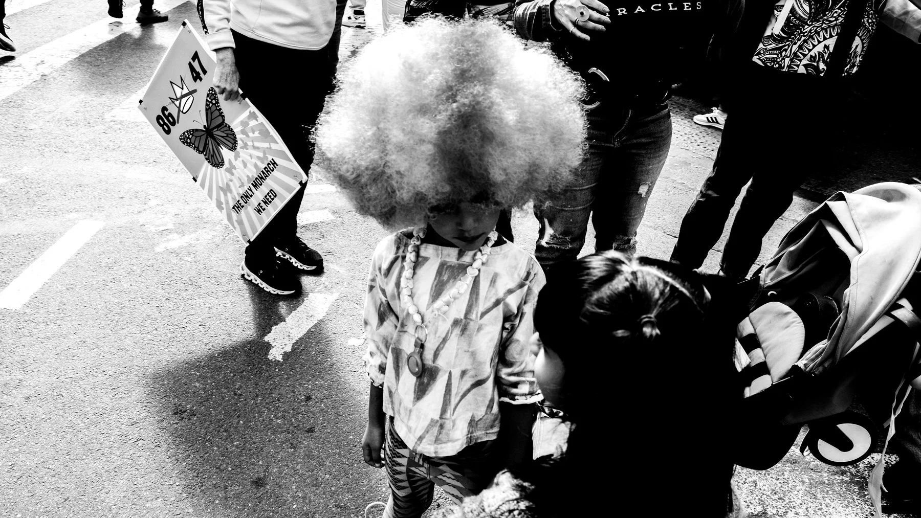 Two children, one with curly bright hair and a necklace, are looking down and talking to each other on a street, with several adults around them and a stroller nearby.