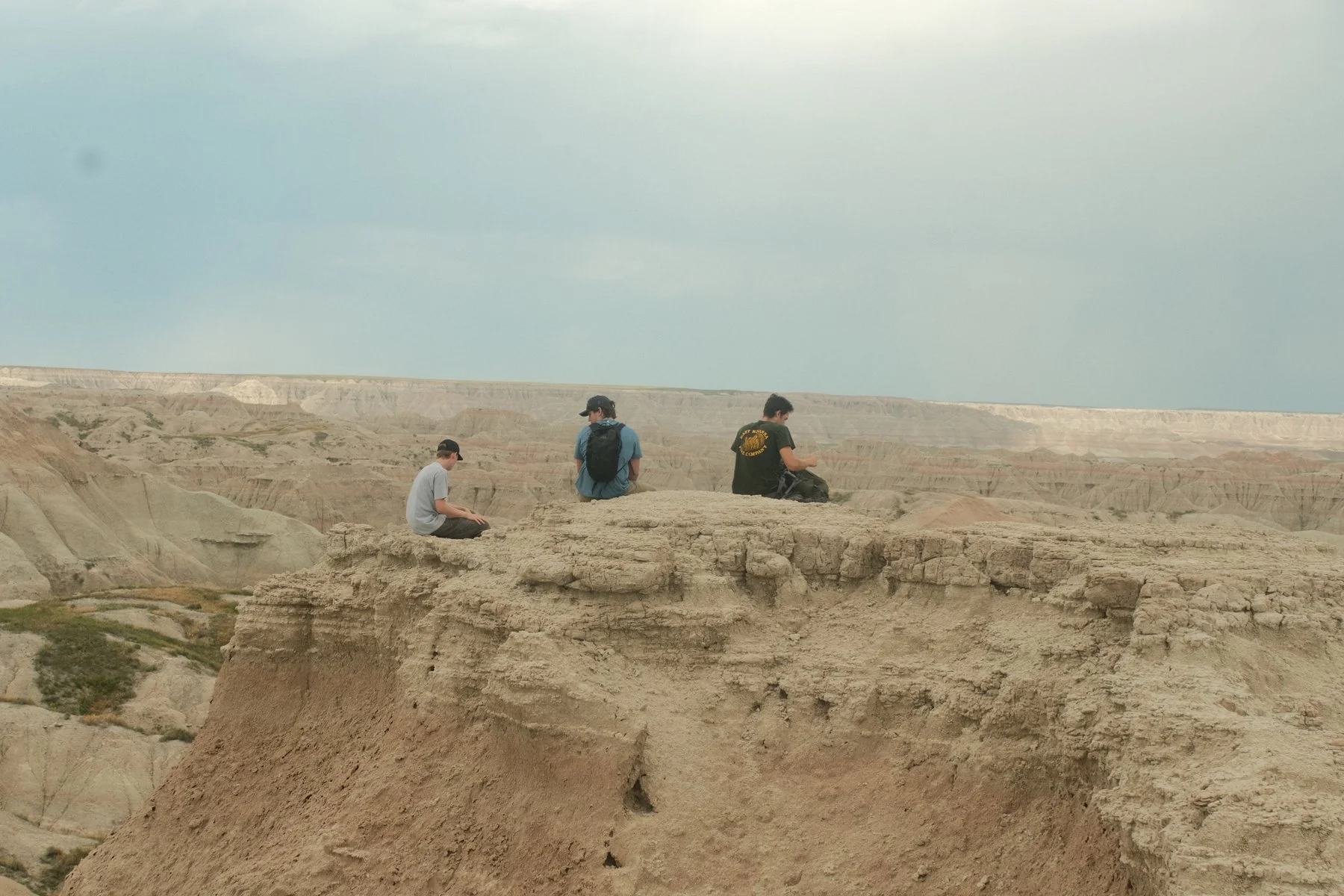 Three people sitting and relaxing on a rocky edge of a canyon, with vast eroded land formations in the background under a cloudy sky.