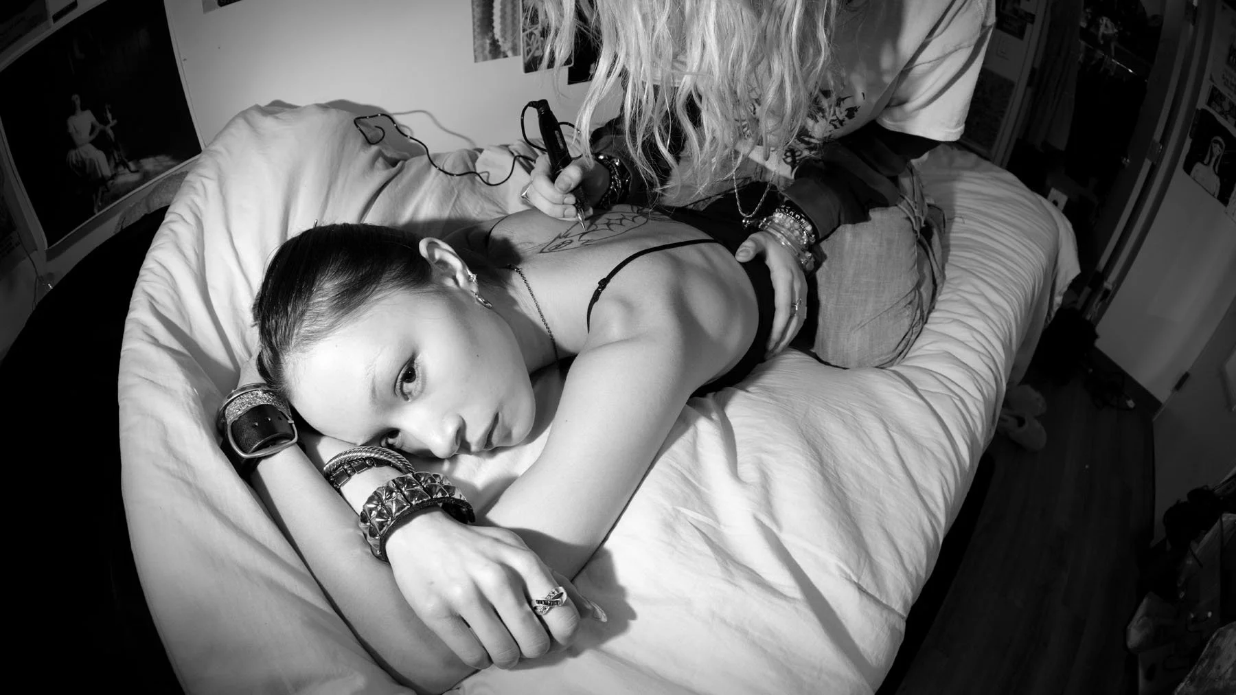 A young woman with multiple bracelets and earrings lying on a bed, looking at the camera. She is being tattooed by an artist with curly hair, who is standing over her, drawing or tattooing on her back.