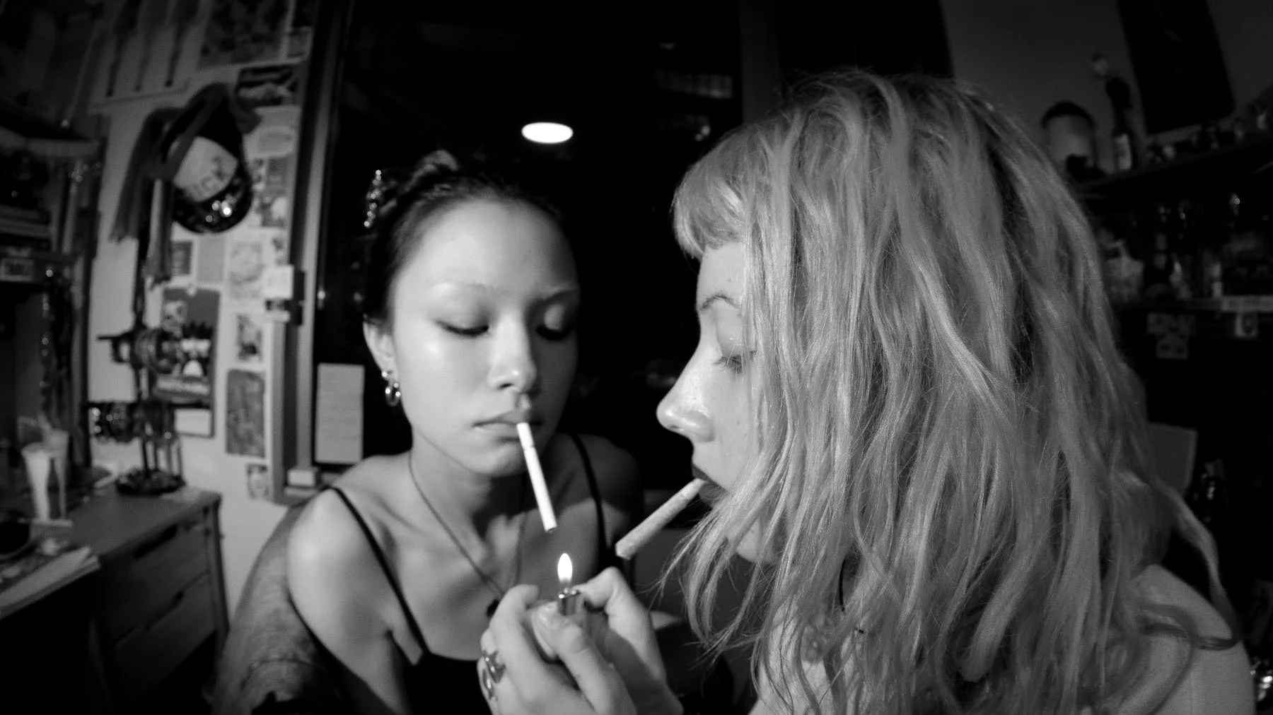 Two women with cigarettes in their mouths, sharing a lighter in a cluttered room.
