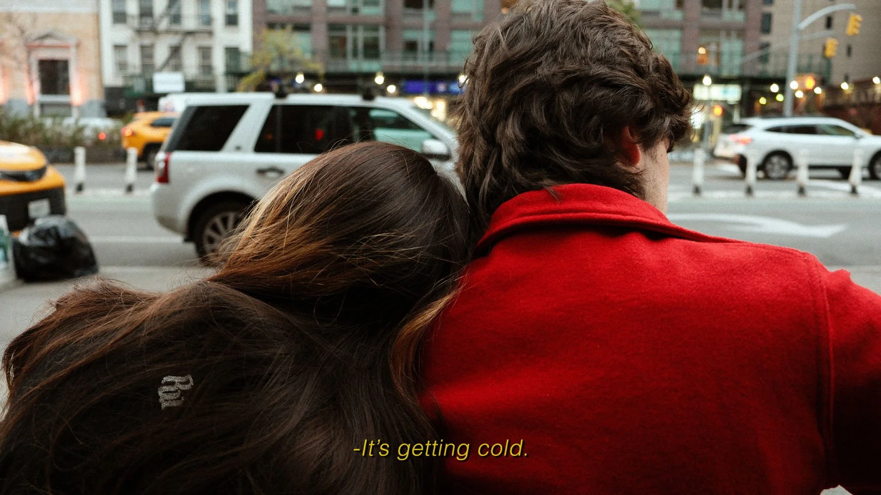 A woman with long dark hair leaning on a man with curly brown hair, wearing a red jacket, sitting on a bench in an urban setting. Cars and buildings are visible in the background, and the subtitle reads, 'It's getting cold.'