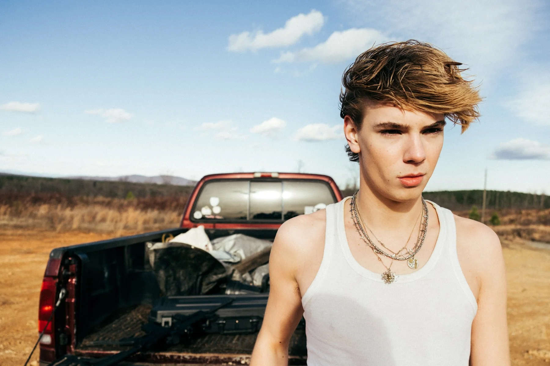 Young person with short, tousled hair wearing a white tank top and layered necklaces outdoors near a red pickup truck loaded with gear, in a rural landscape under a partly cloudy sky.