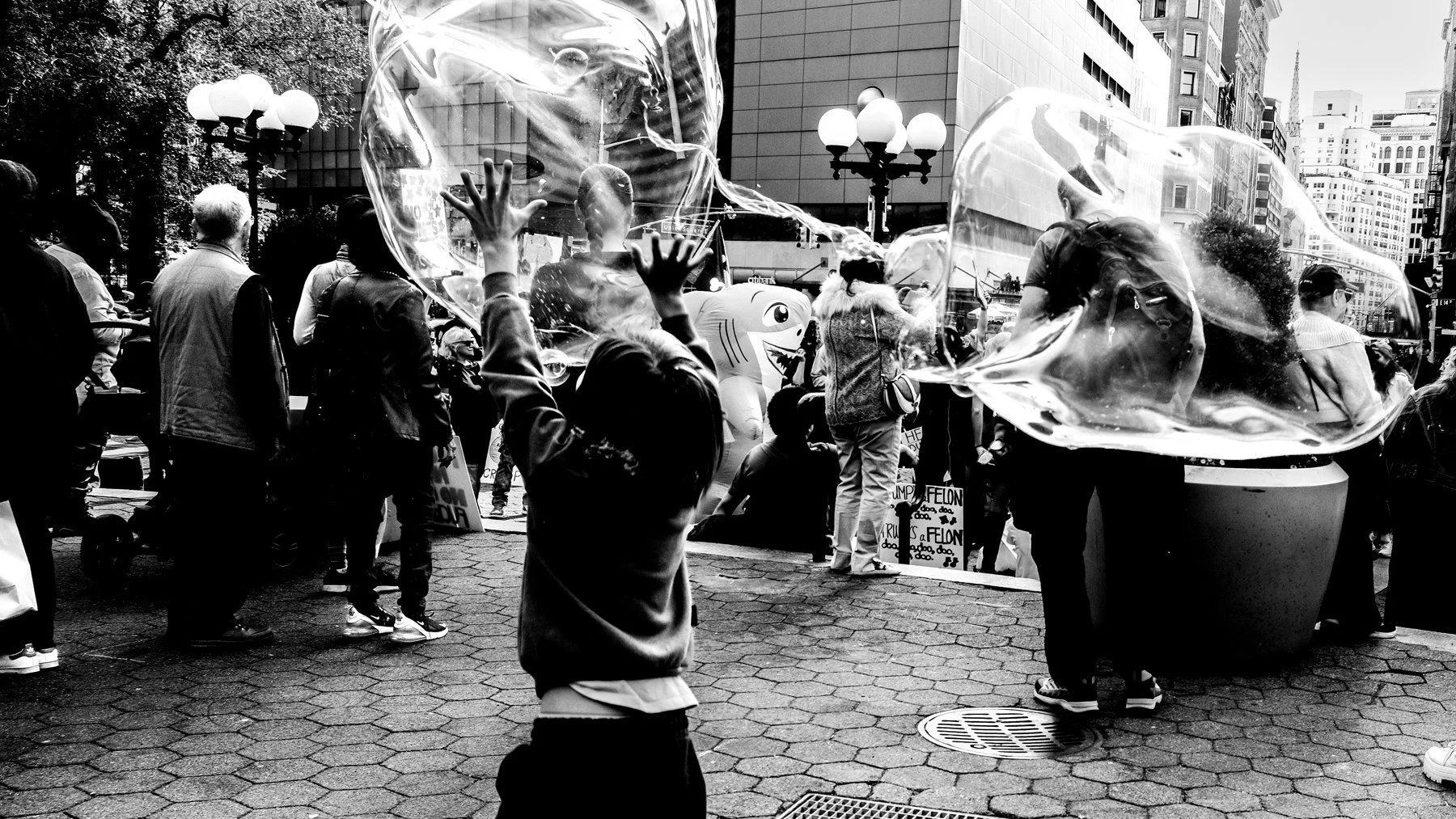 A black and white photo of a crowded city outdoor protest or rally. People are gathered, some holding signs. Two large soap bubbles are in the foreground, with children and adults around, some trying to touch or look at the bubbles. Tall buildings an