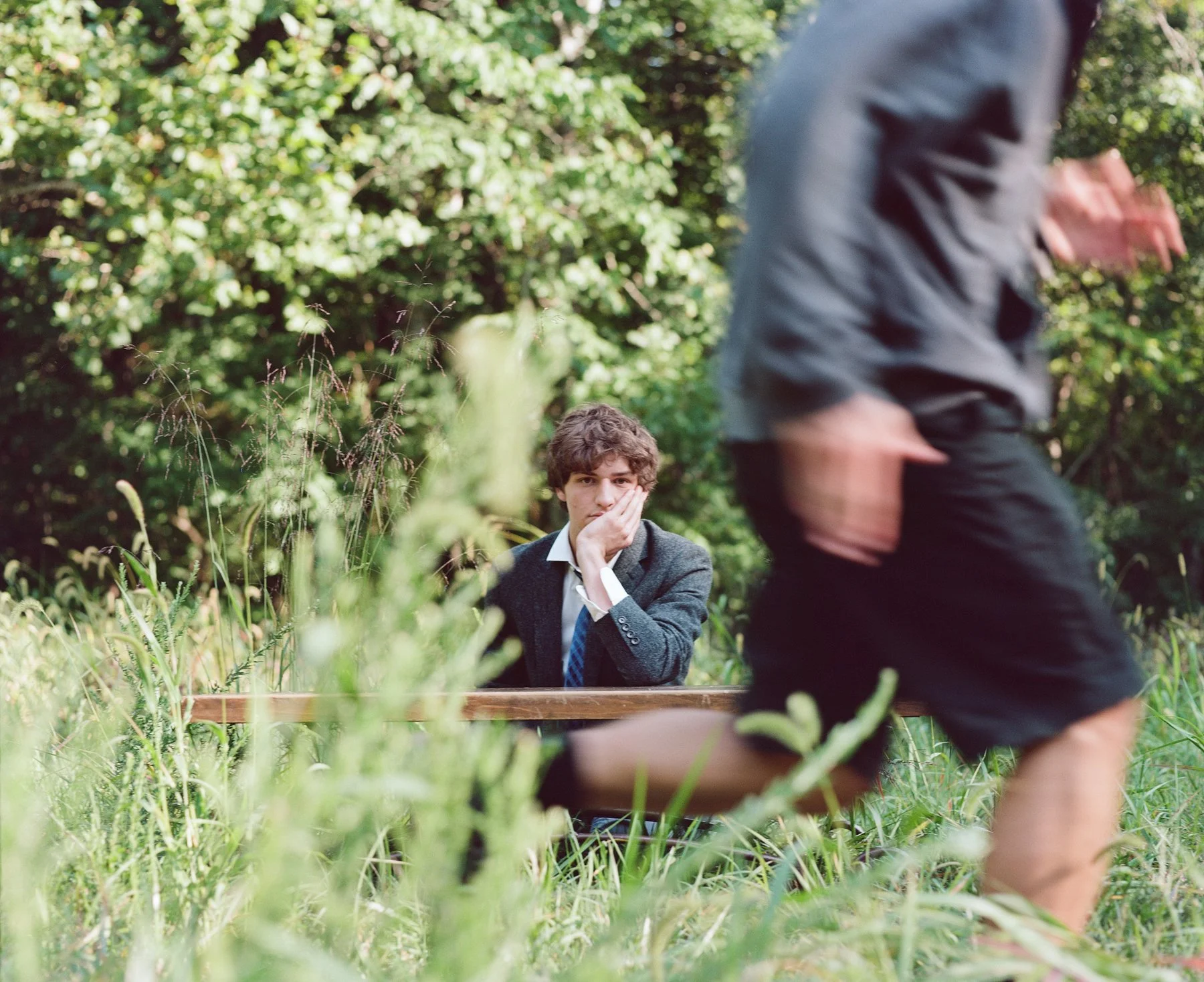 A young man in a suit and tie sitting on a bench in a grassy, wooded area, with his hand on his face, looking contemplative. A man wearing shorts and a jacket is walking past in the foreground, slightly out of focus.