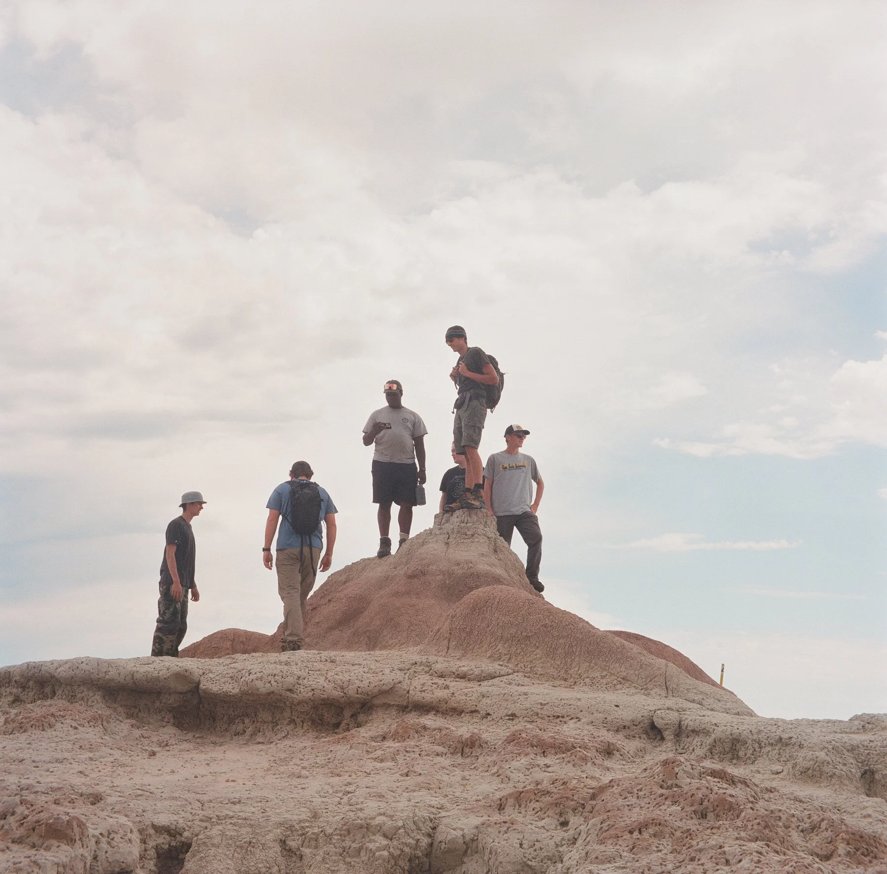 Group of six people standing and walking on a sandy hill with clouds in the sky.