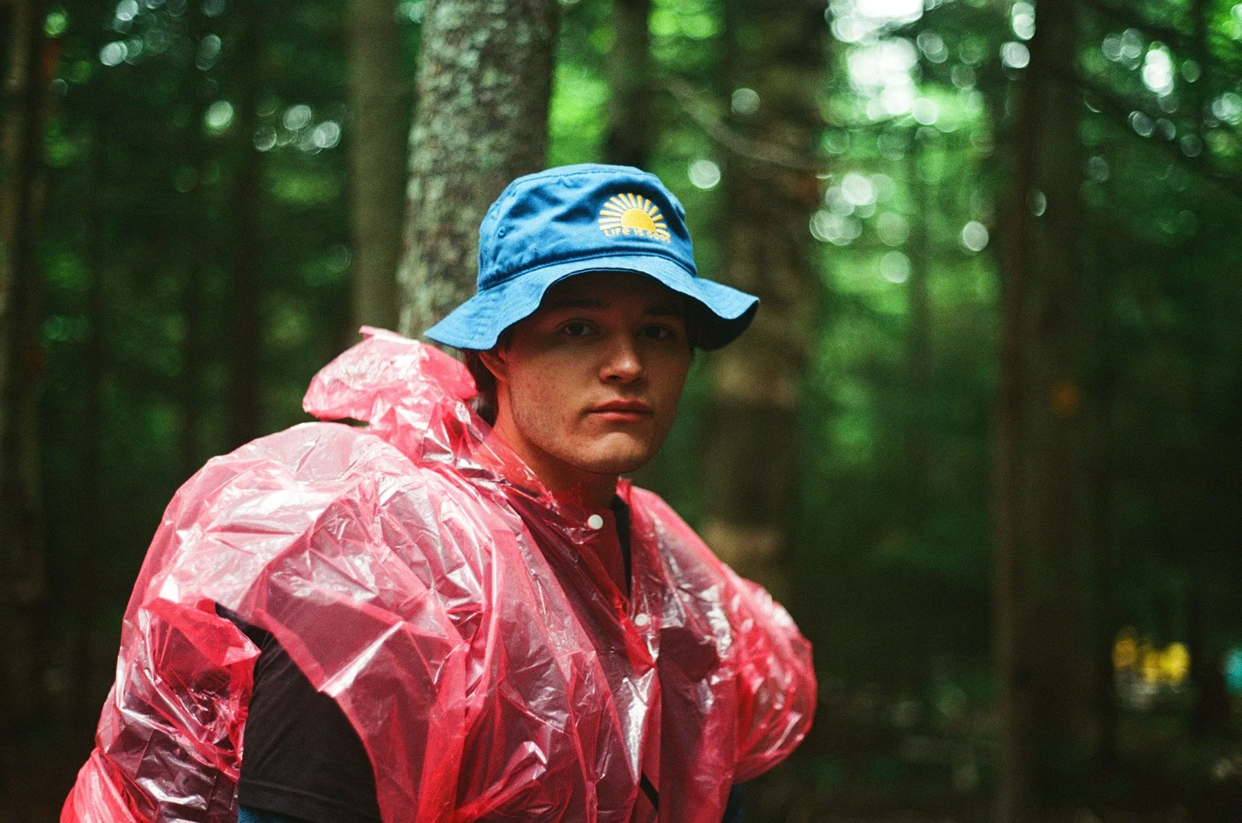 A young man wearing a blue bucket hat with a yellow sun logo and a red waterproof jacket standing in a lush green forest.