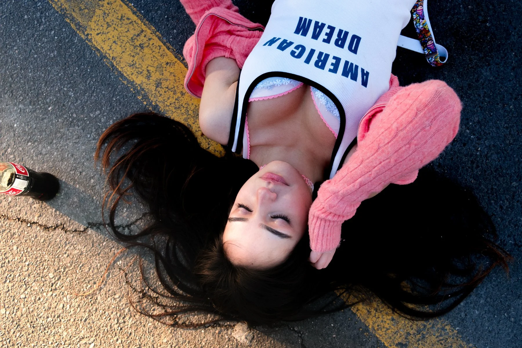 A young woman with long dark hair lying on the pavement, eyes closed, wearing a white tank top with blue text, a pink cardigan, and a choker necklace. There is a Coca-Cola bottle nearby.