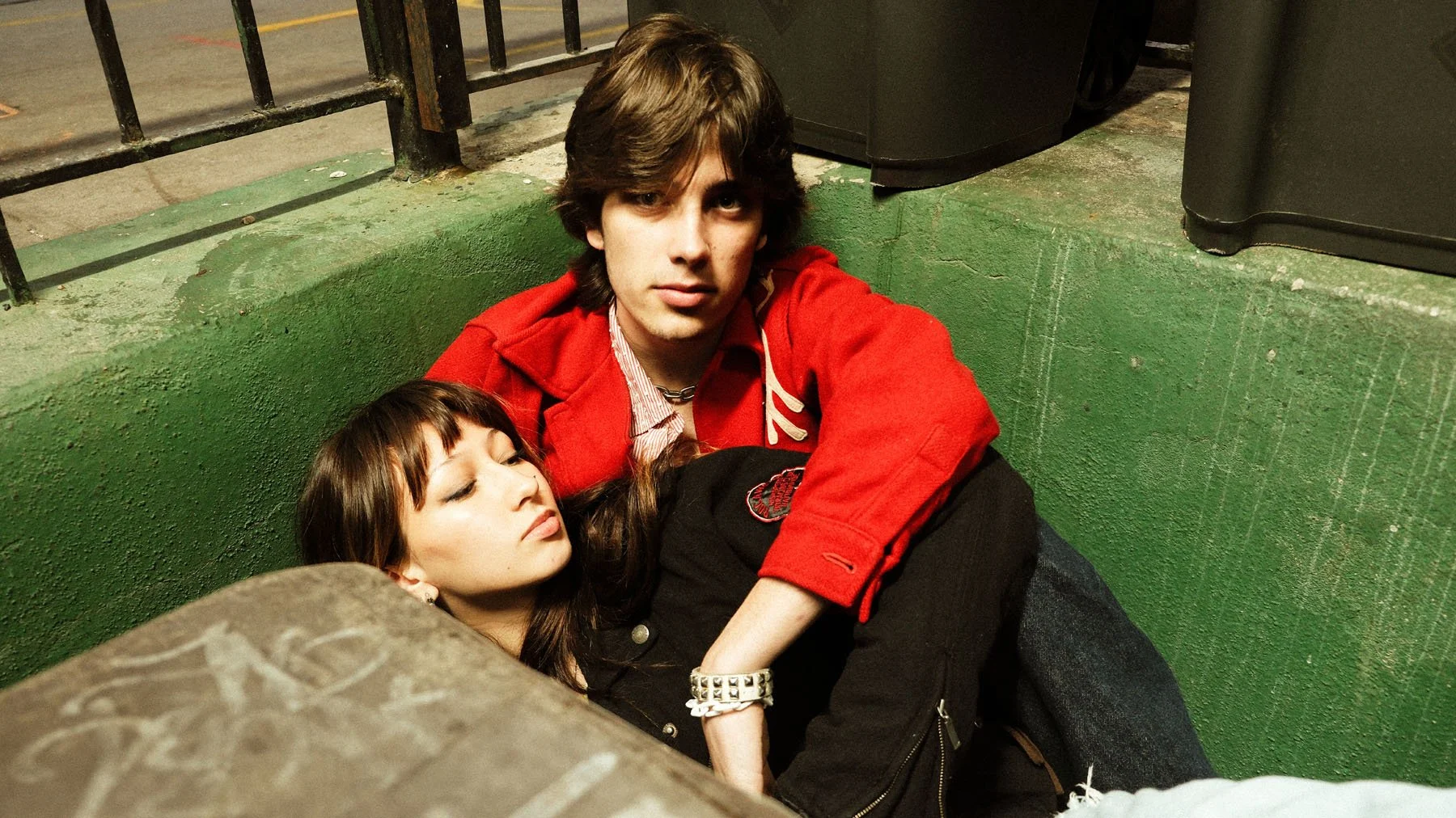 A young man and woman lying on the ground in an urban setting with a green wall, black railing, and trash bins nearby. The woman has long brown hair and appears to be unconscious or resting, while the man has medium-length brown hair and looks direct