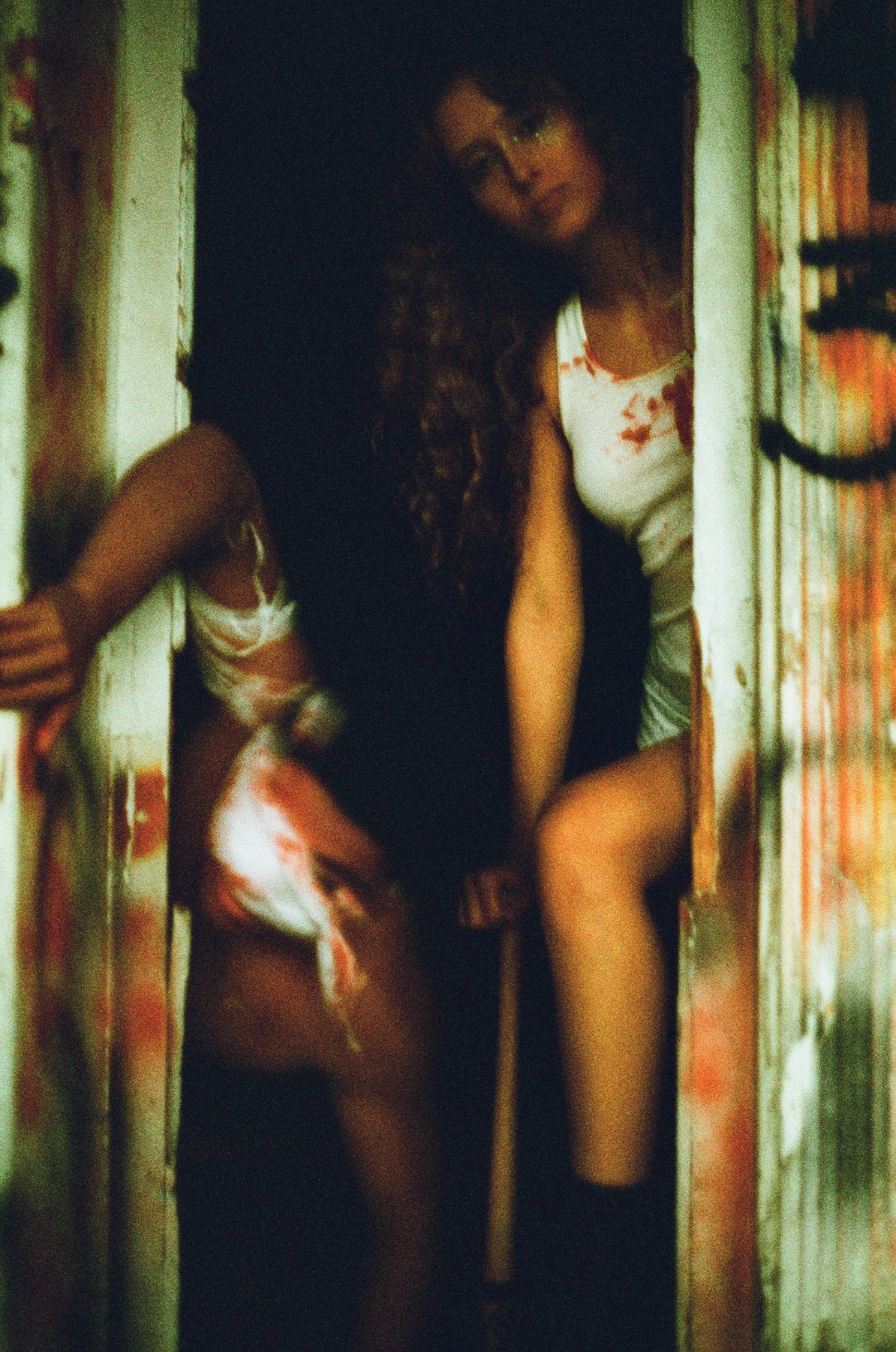 Two women peeking through a small opening in a weathered, graffiti-covered wall, wearing casual attire with bloodstains, holding an object, with a dark background.