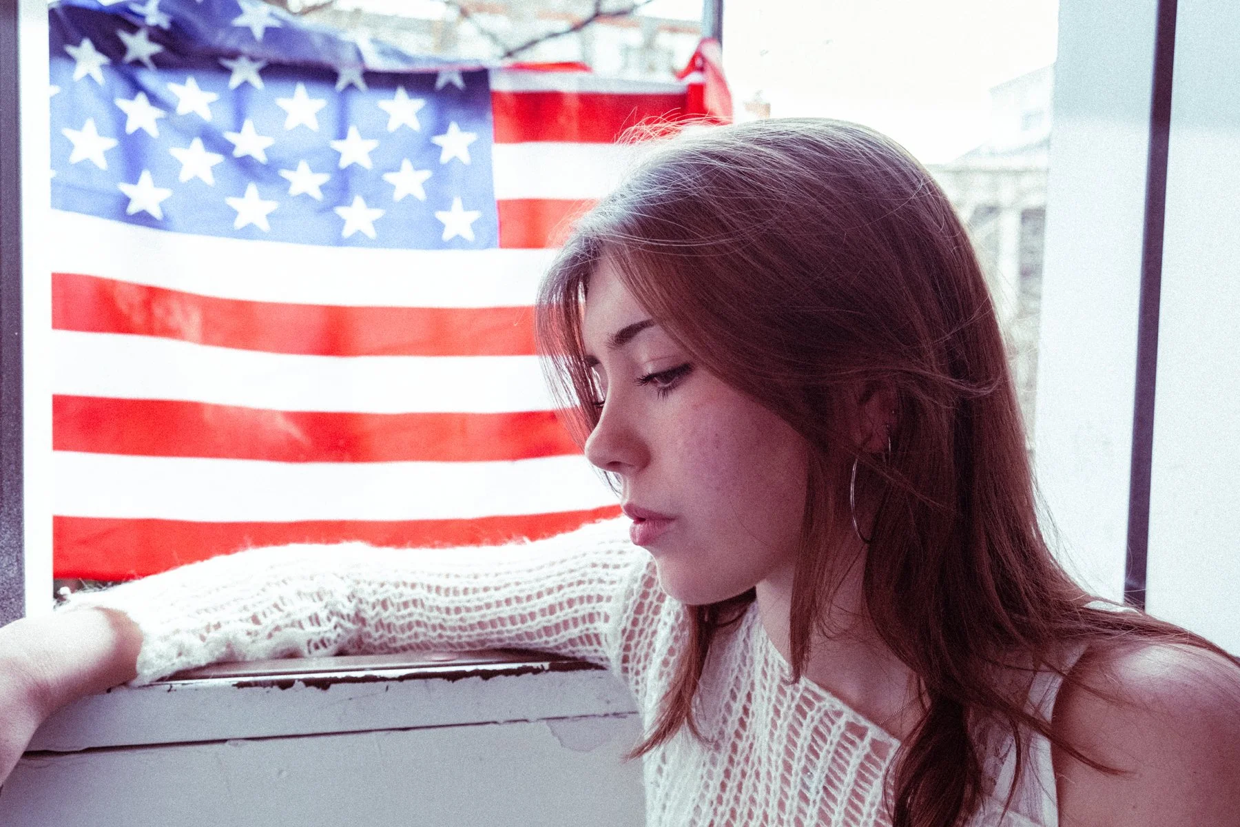 A young woman with reddish-brown hair wearing a white sweater sitting by a window with an American flag in the background.