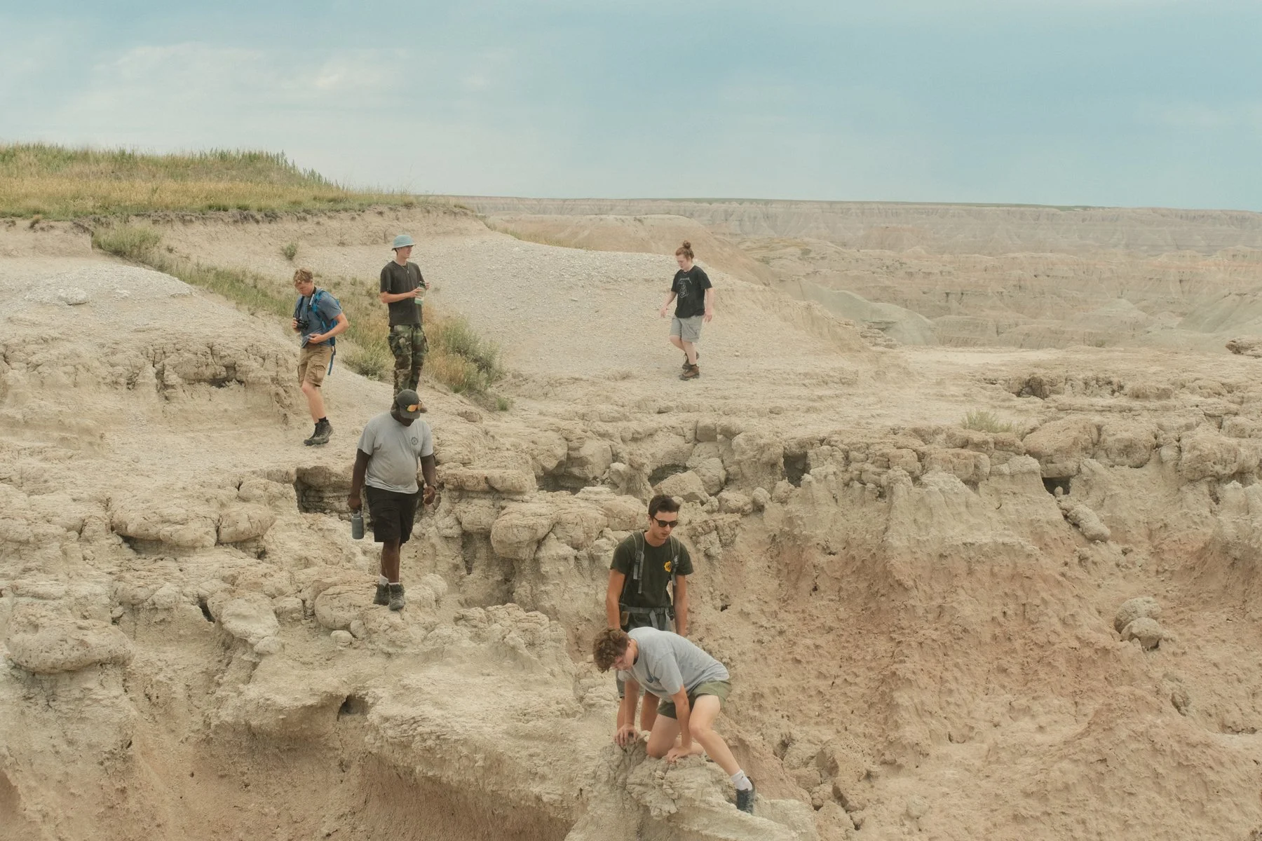 A group of seven people exploring a desert landscape with eroded canyon formations, some with cameras and backpacks, some climbing or walking on the rocky terrain.