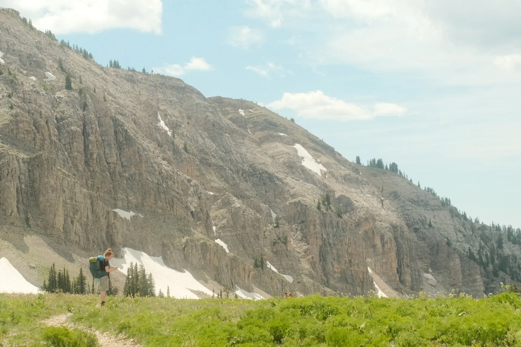 A hiker standing on a grassy trail in front of a mountain with patches of snow and scattered trees under a partly cloudy sky.