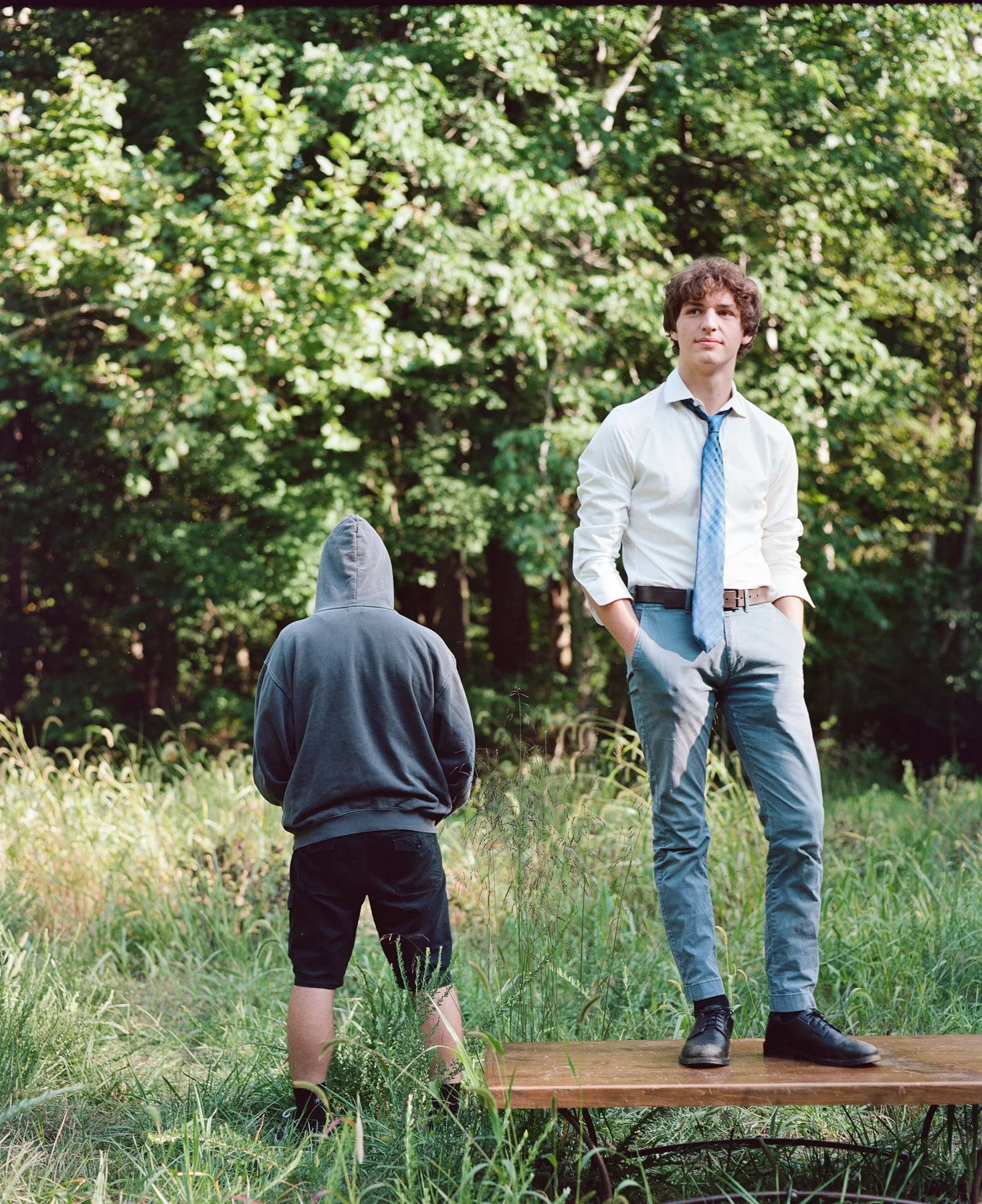 A young man in formal attire standing on a wooden platform outdoors, with a hooded figure in casual clothing facing away in the grass and trees.