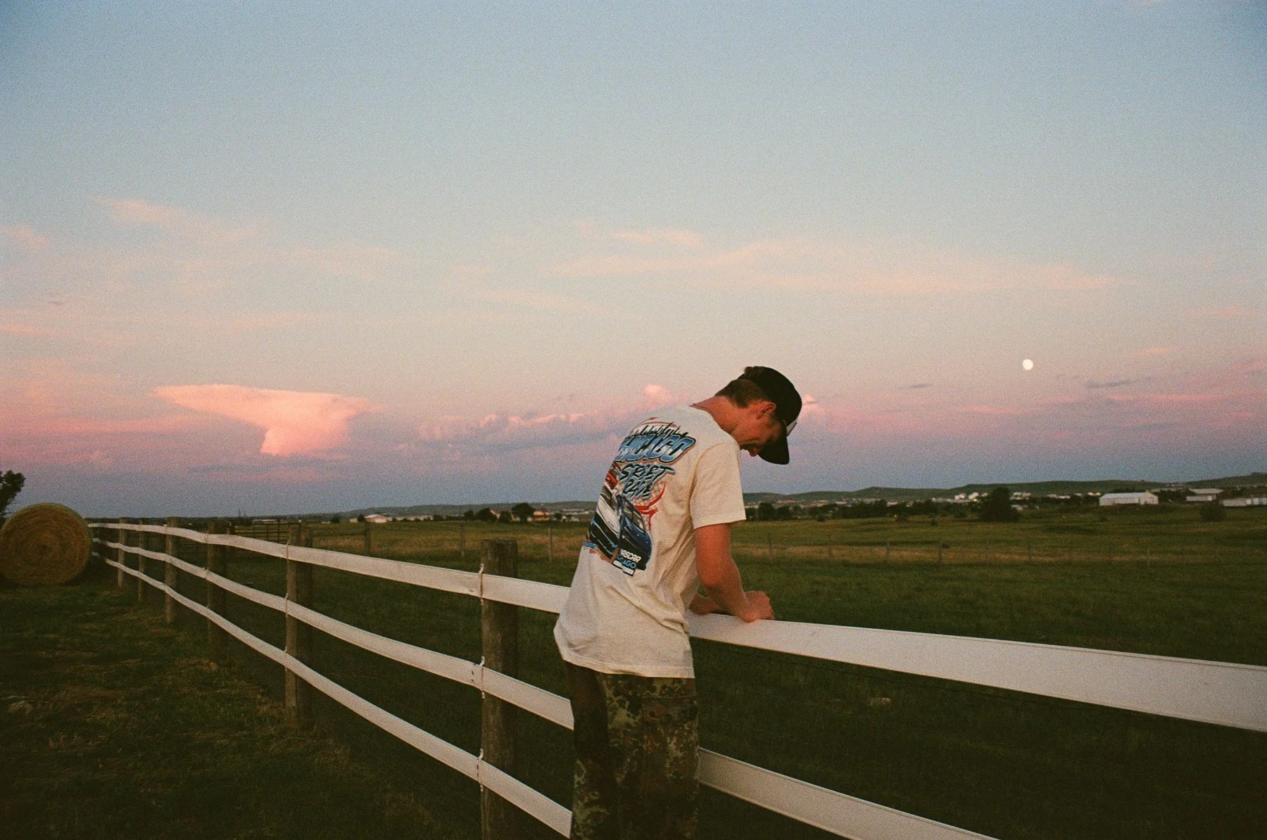 A young man with a dark baseball cap, a graphic T-shirt, and camouflage pants leaning on a white wooden fence in a rural field during sunset, with the moon visible in the sky.