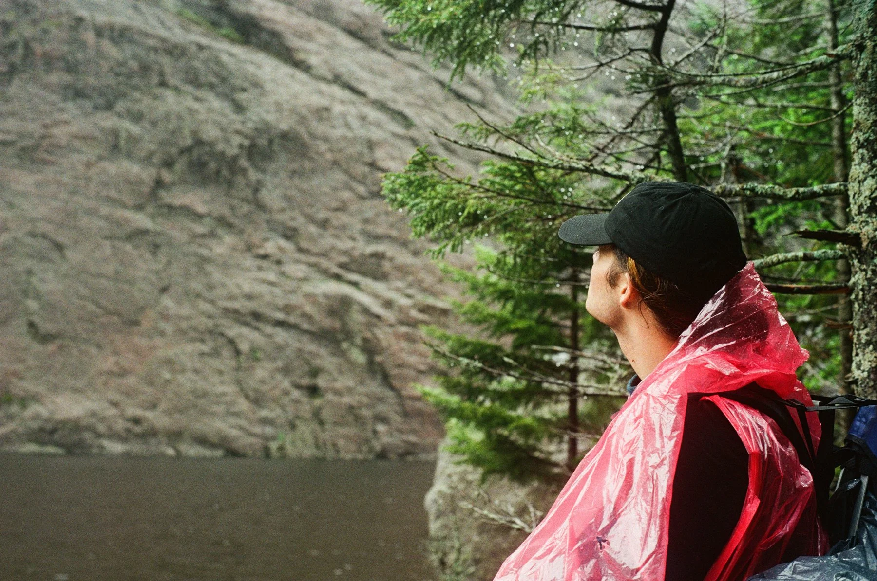 Person wearing a black cap and red rain poncho sitting on a rock near a river, surrounded by green trees in a forested area.