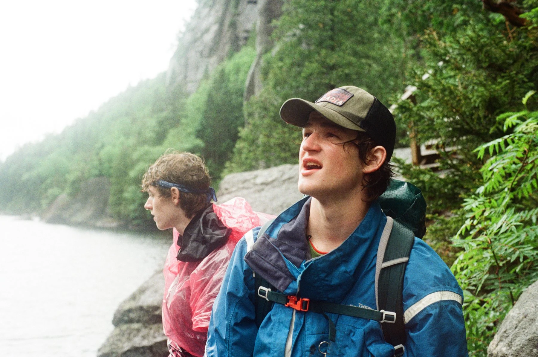 Two young men in outdoor rain gear standing by a rocky lakeshore with green trees and cliffs in the background, one looking towards the water with a concerned expression.