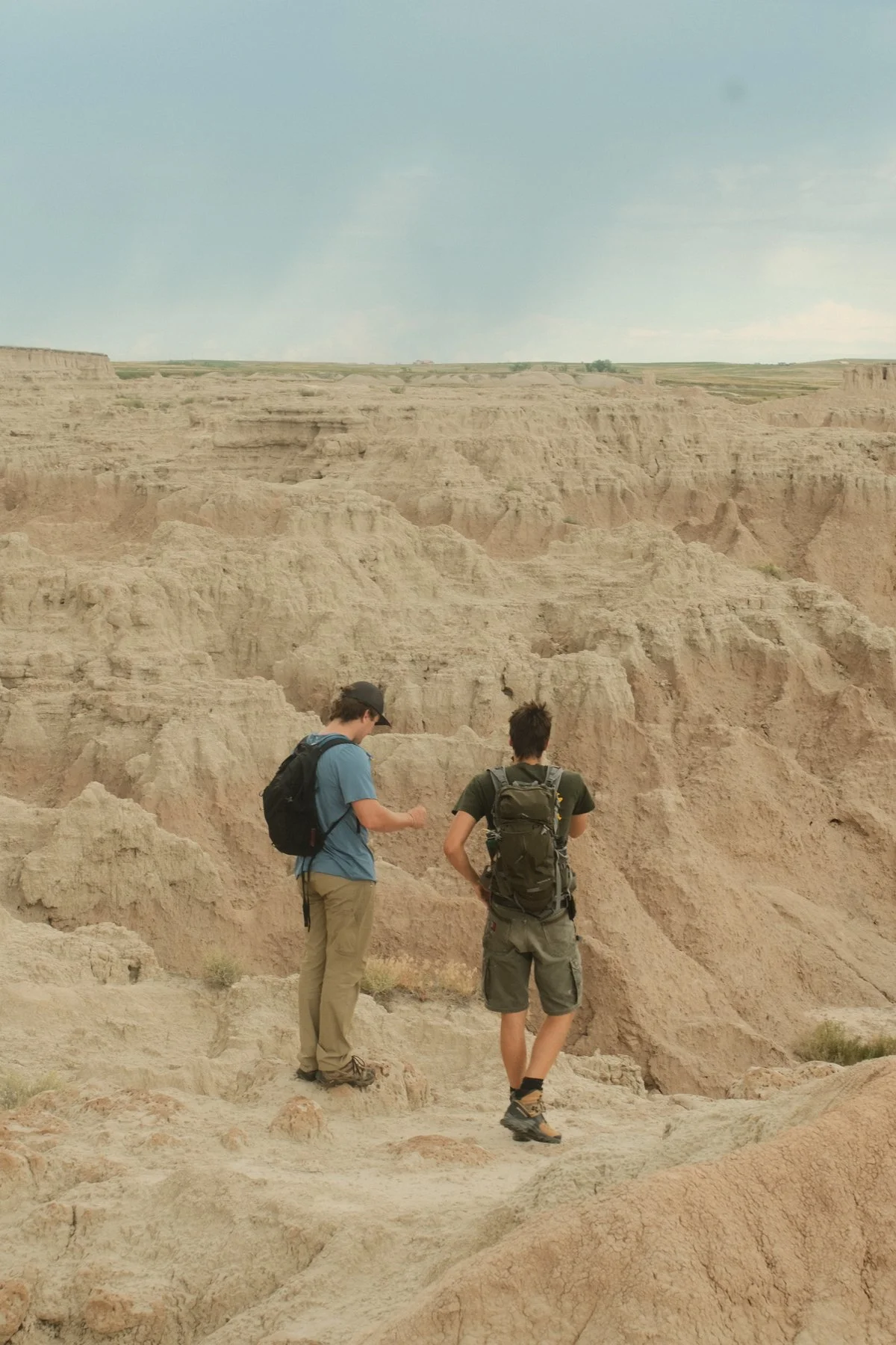 Two young men with backpacks hiking in a desert canyon, looking at the rugged, light-colored rock formations under a partly cloudy sky.