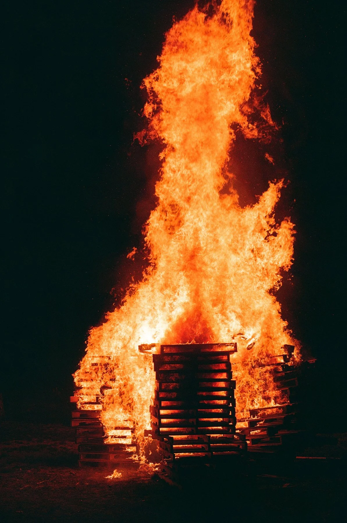 Large bonfire with flames rising high into the night sky, surrounded by wooden pallets.