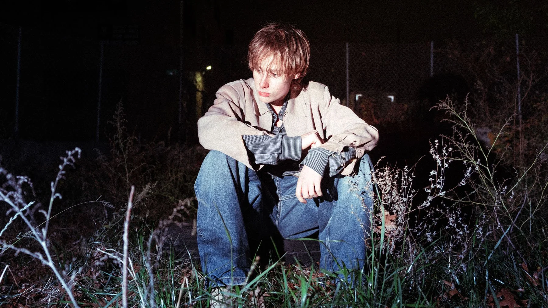 A young person with light brown hair sits alone outdoors at night, surrounded by tall grass and weeds. They are wearing a beige jacket, a plaid shirt, and jeans, looking contemplative with their arms resting on their knee.