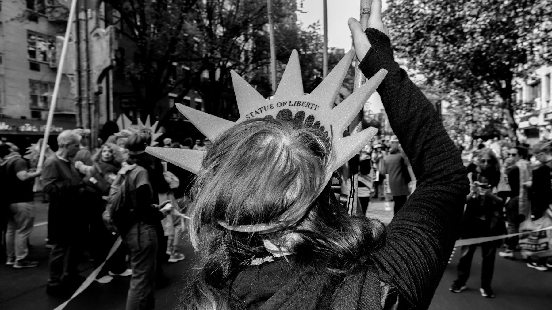 A person wearing a Statue of Liberty crown at a parade, with a crowd of people and trees in the background.
