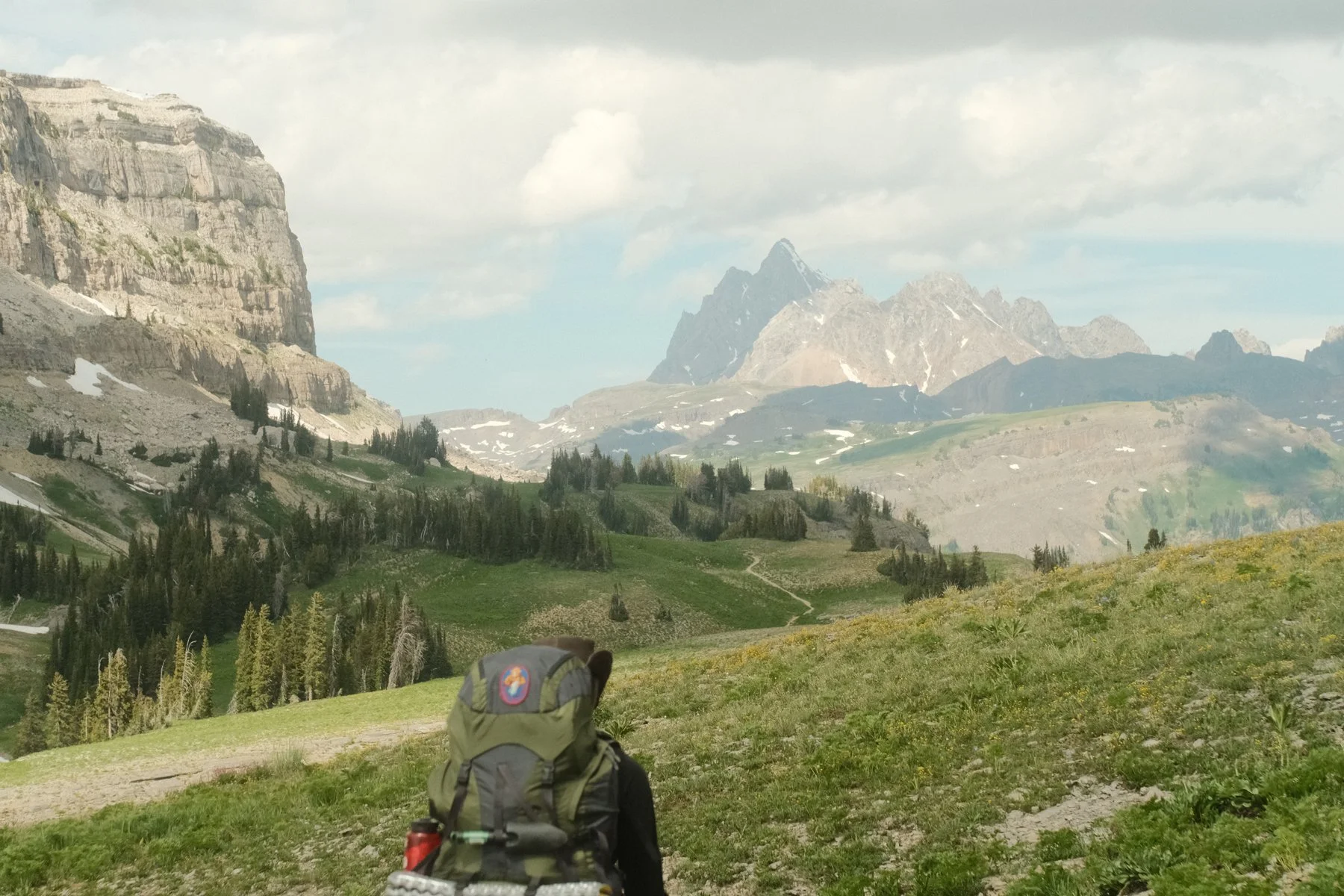 Hiker with a backpack walking through a lush green mountain landscape with rocky peaks and scattered trees under a partly cloudy sky.
