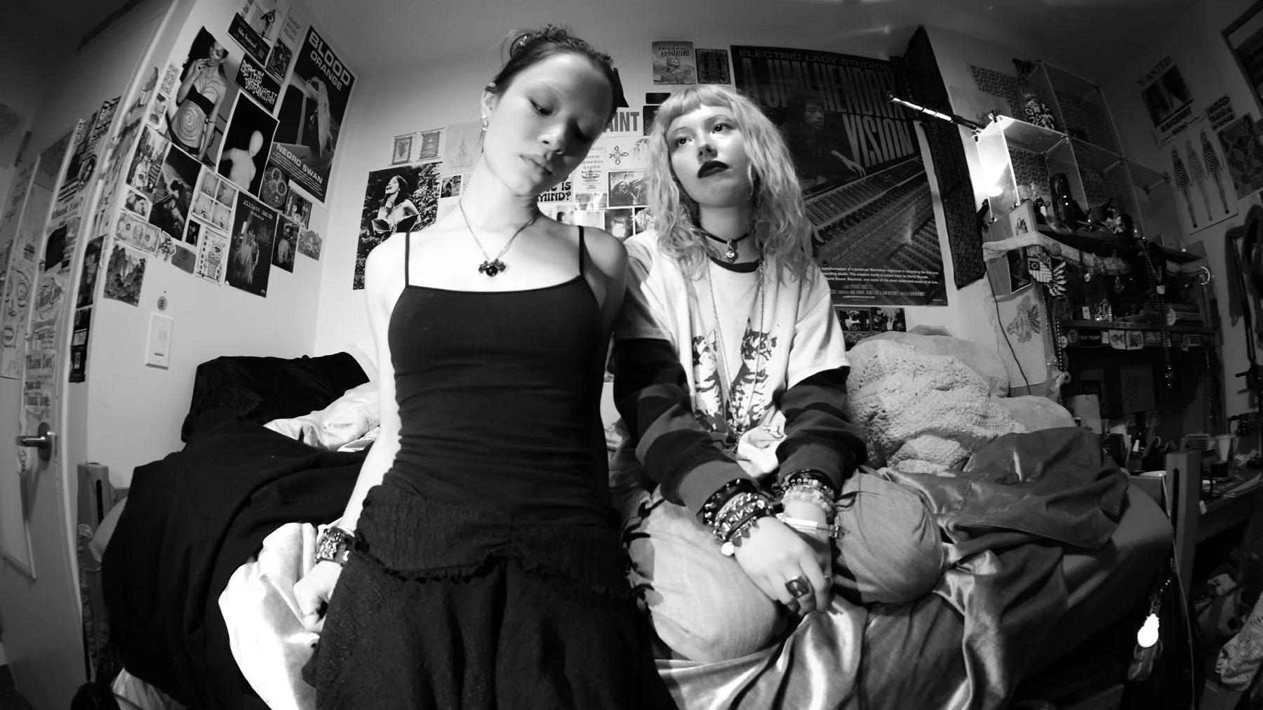 Two young women sit on a bed in a cluttered room filled with posters and artwork on the walls. The woman on the left wears a black spaghetti strap top and looks down, while the woman on the right has curly hair, dark lipstick, and wears layered cloth