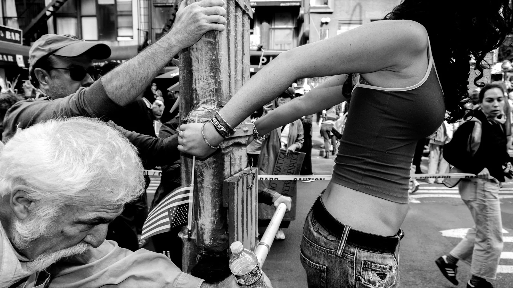 A woman with tattoos on her arms wearing a tank top and jeans pushes against a police barrier in a crowded street with several pedestrians and a protest sign visible in the background.