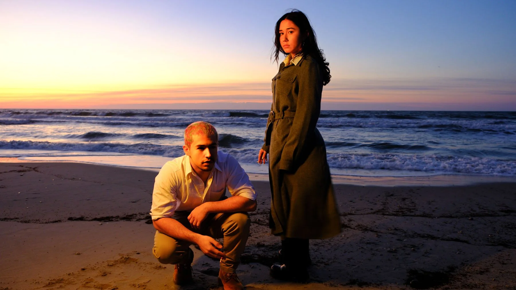 A man squatting on the beach with a woman standing nearby, both during sunset with the ocean in the background.