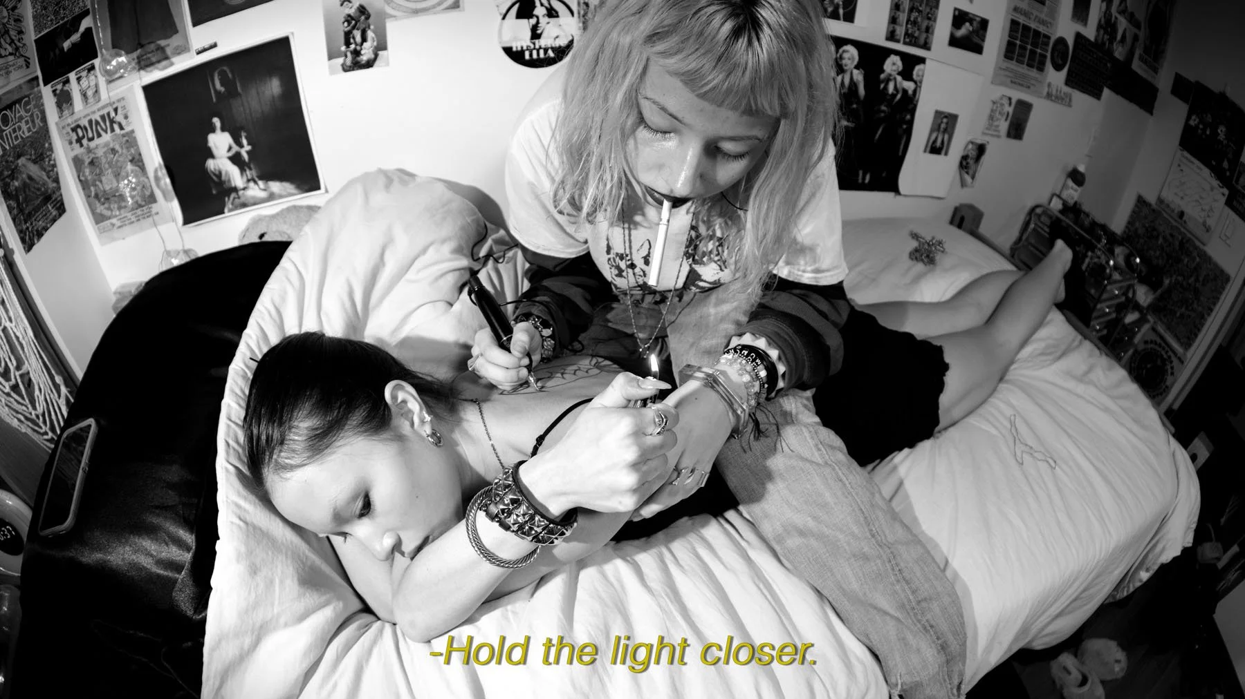 A woman with blonde hair and tattoos on her arm is tattooing a young woman lying on her stomach on a bed. The young woman has dark hair, earrings, and bracelets, and is resting her head on her arm. The room has various posters and pictures on the wal