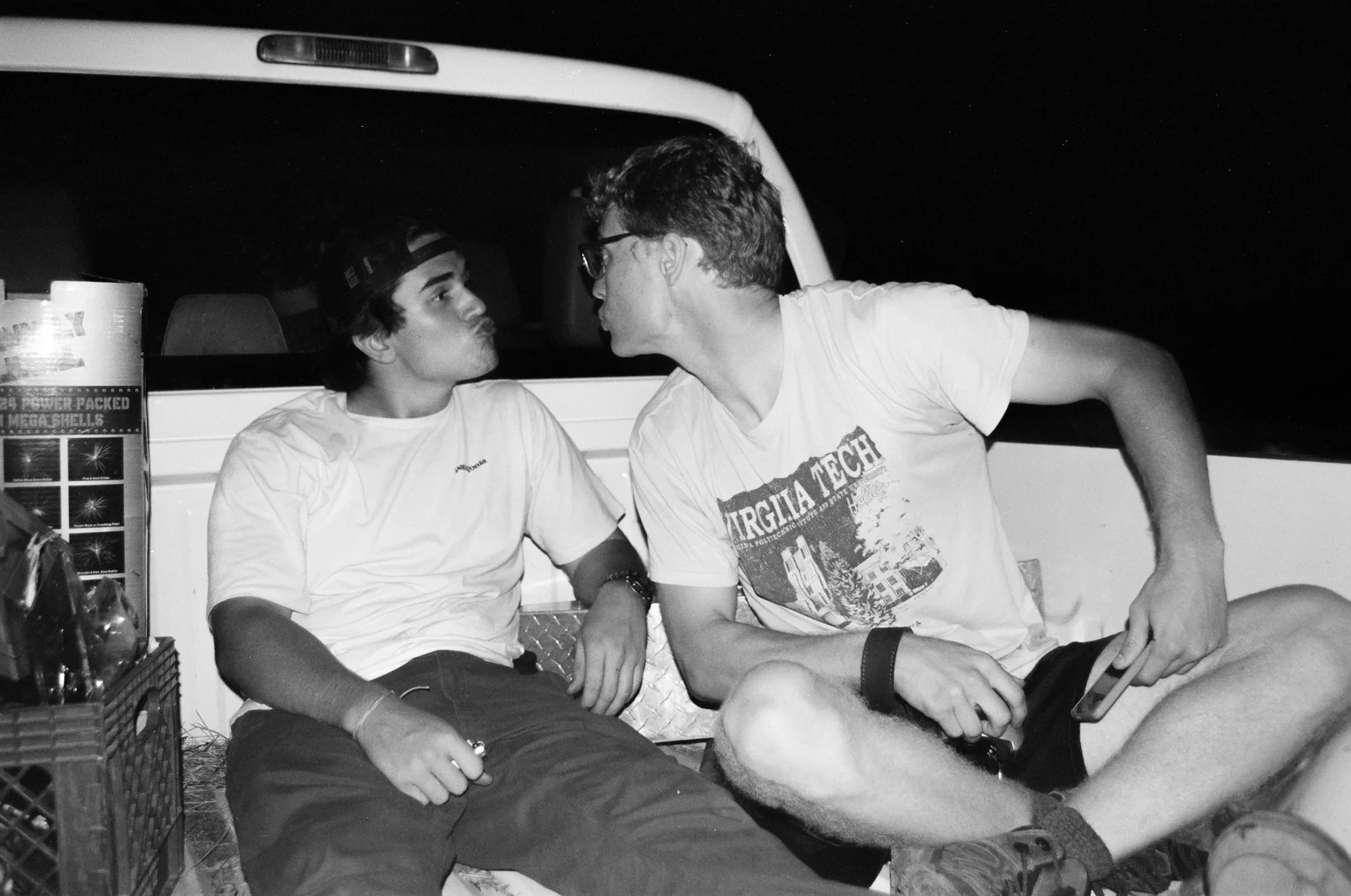 Two young men sitting in the bed of a pickup truck at night, leaning towards each other with puckered lips as if about to kiss.