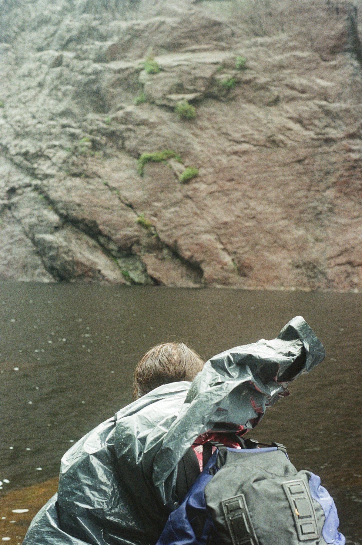 A person with brown hair facing a body of water with a rocky cliffside in the background, wearing a backpack and a rain jacket.
