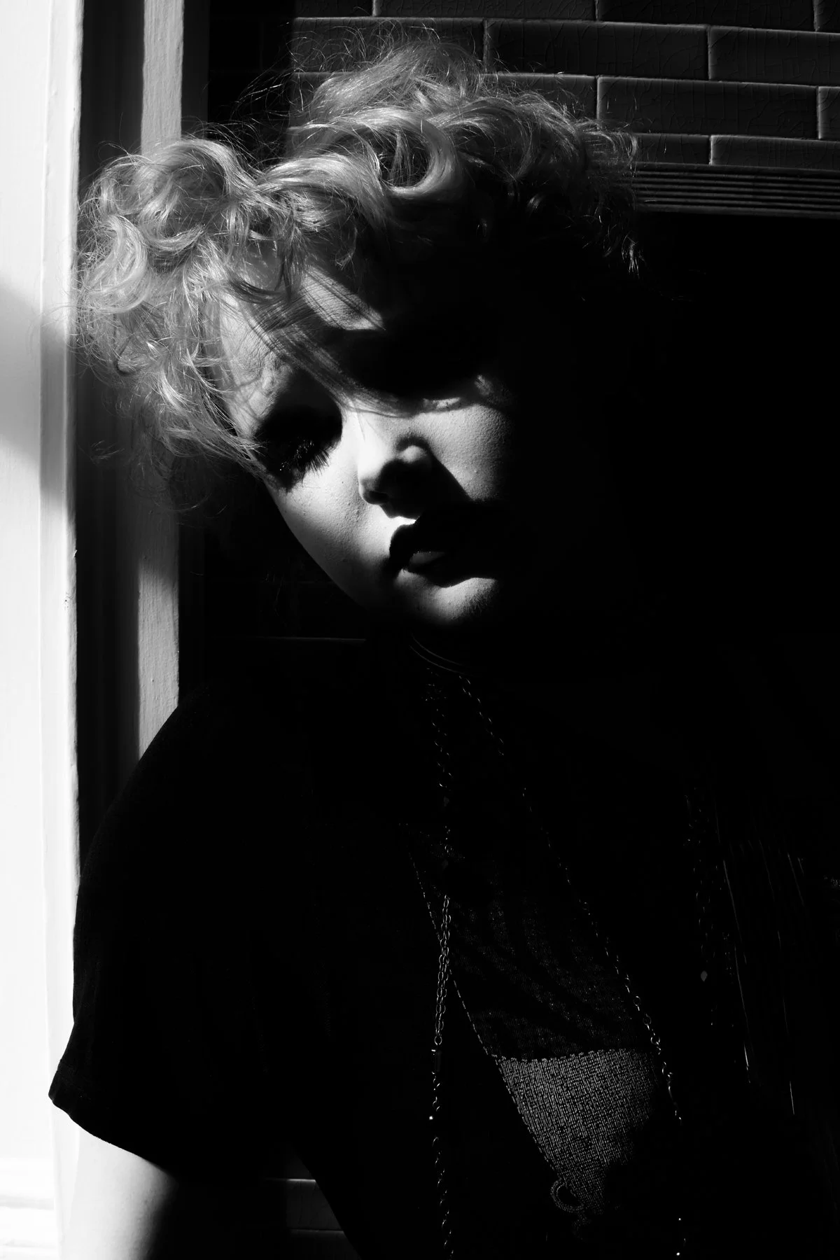 A black and white photograph of a woman with curly hair, partially illuminated, with shadows obscuring parts of her face, standing near a wall.