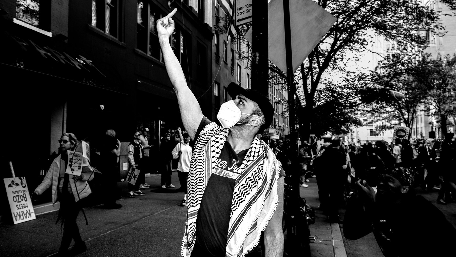 A man wearing a face mask and a keffiyeh scarf is raising his fist in the air during a protest or rally, surrounded by a crowd of people on a city street with trees and buildings.