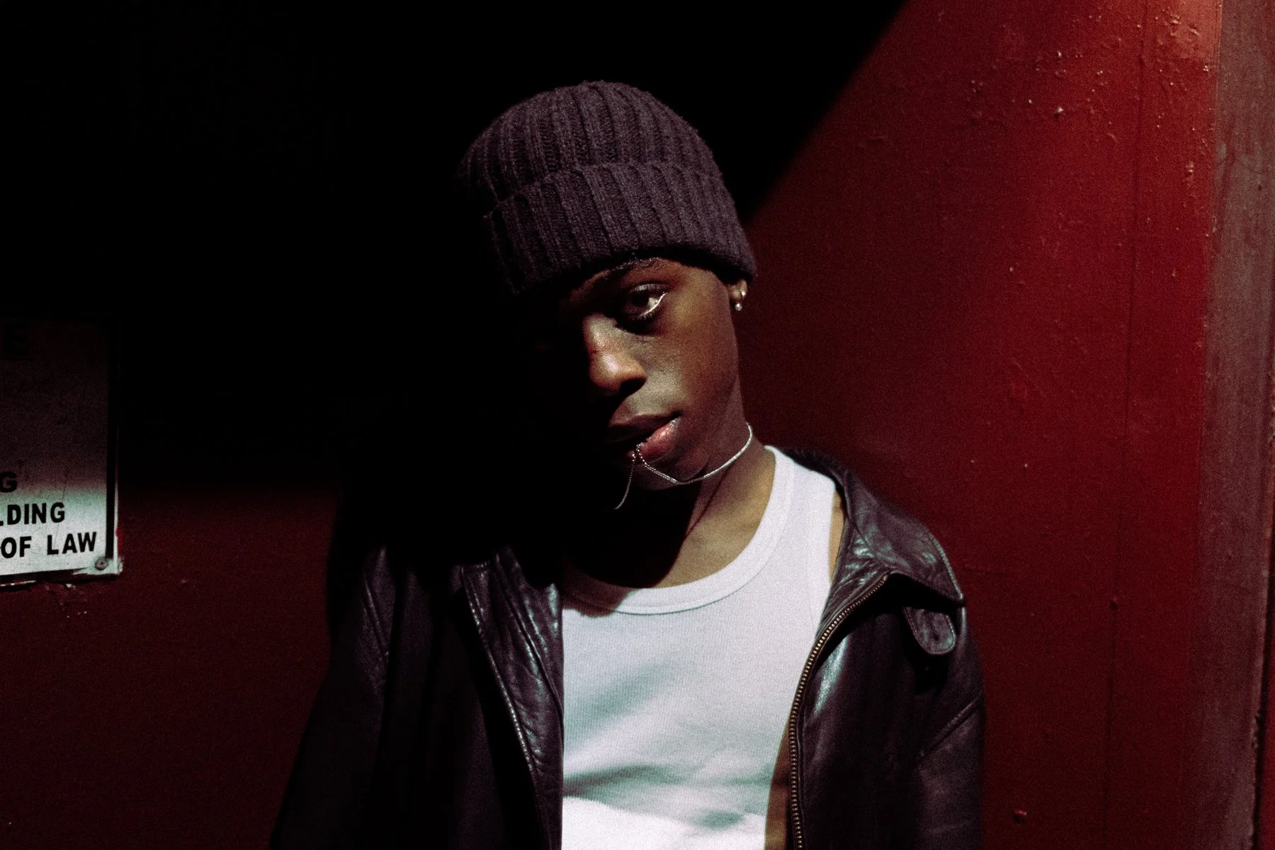 A young man wears a purple beanie, a white t-shirt, and a black leather jacket, standing in a dimly lit environment with a red wall behind him and a shadowed area on the left.